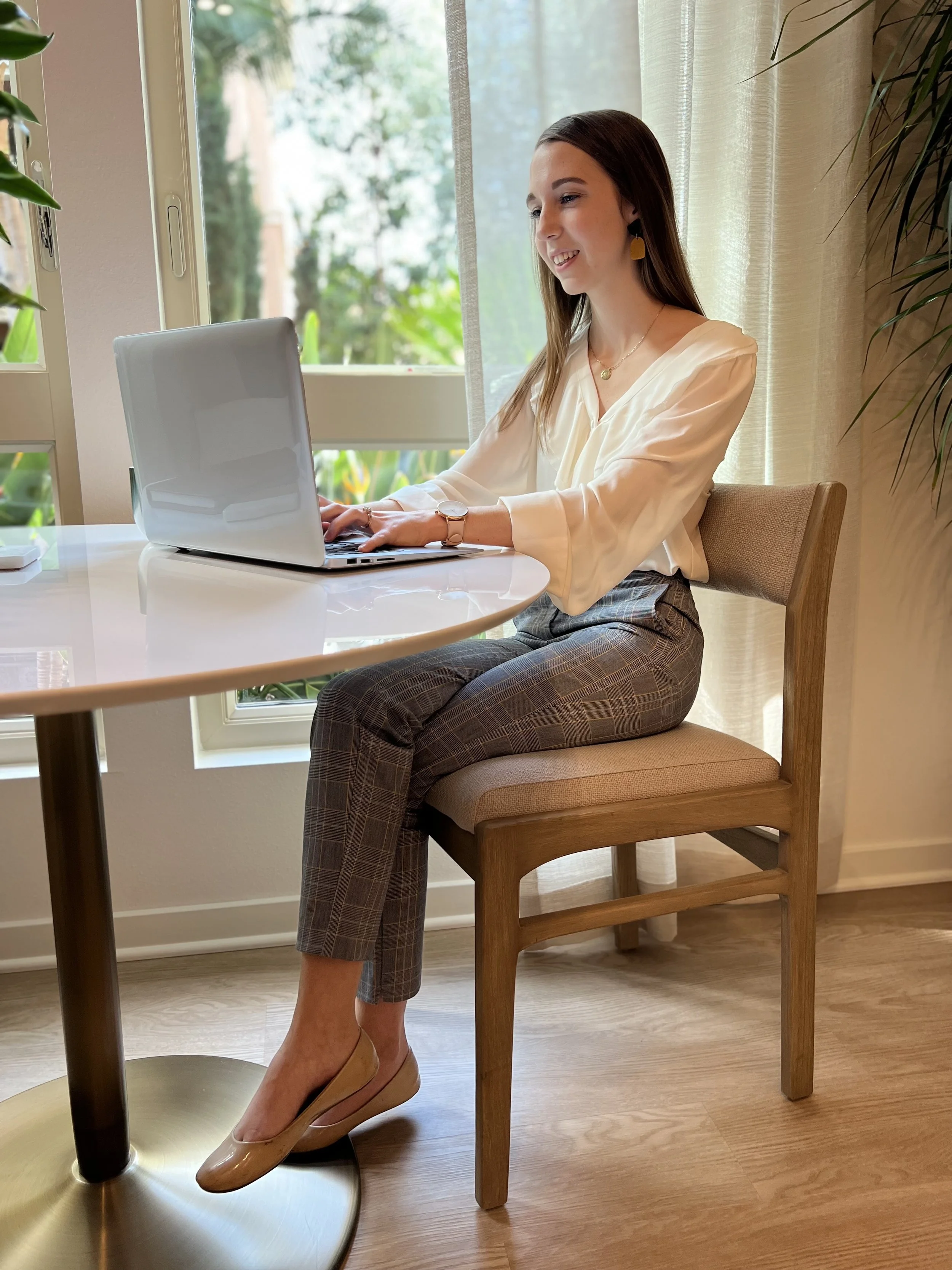 A woman sitting at a dining table using a laptop in a bright room with large windows and curtains, greenery visible outside.