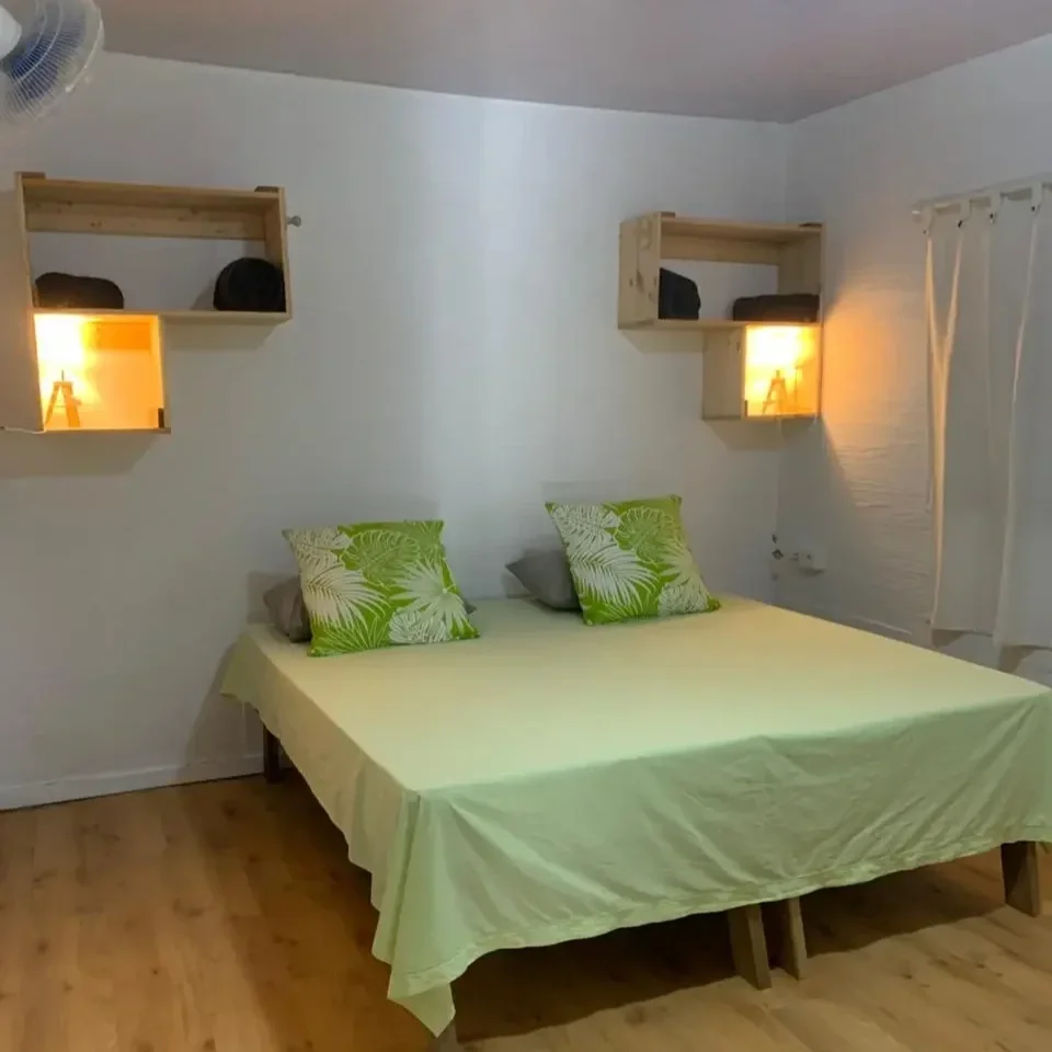 Chambre climatisée avec lit double, deux coussins verts à motifs de feuilles, étagères en bois avec serviettes et couverture, lampes de chevet, rideau blanc sur la fenêtre.