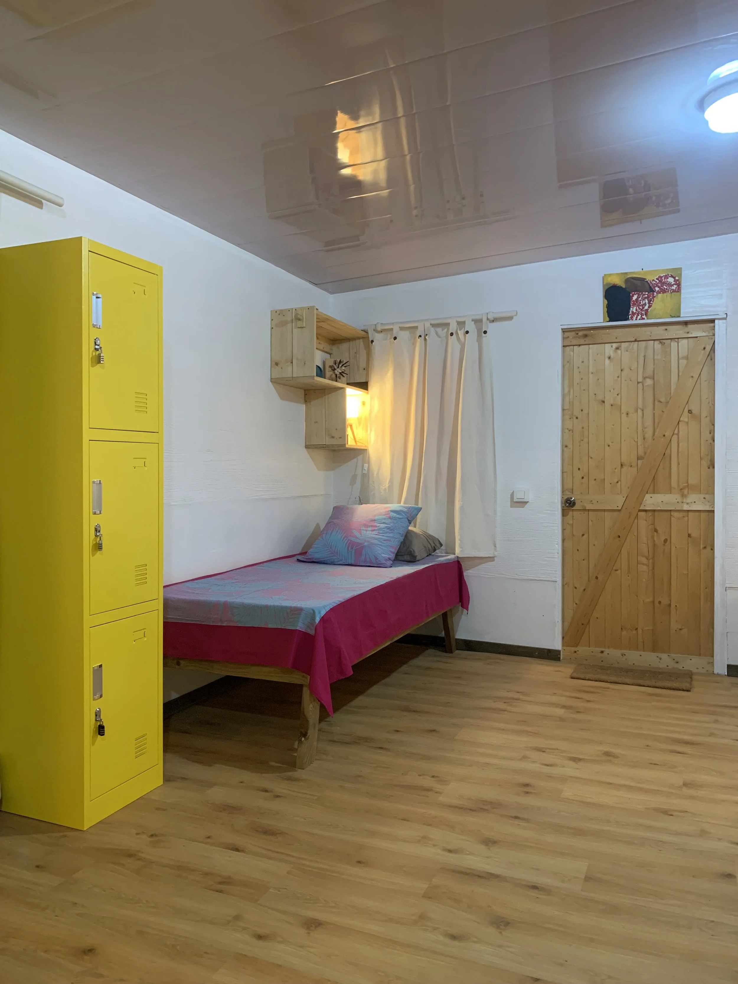 Dortoir mixte avec lit individuel, casier à cadenas jaune, porte en bois, plancher en bois et décoration chaleureuse à Rangiroa.