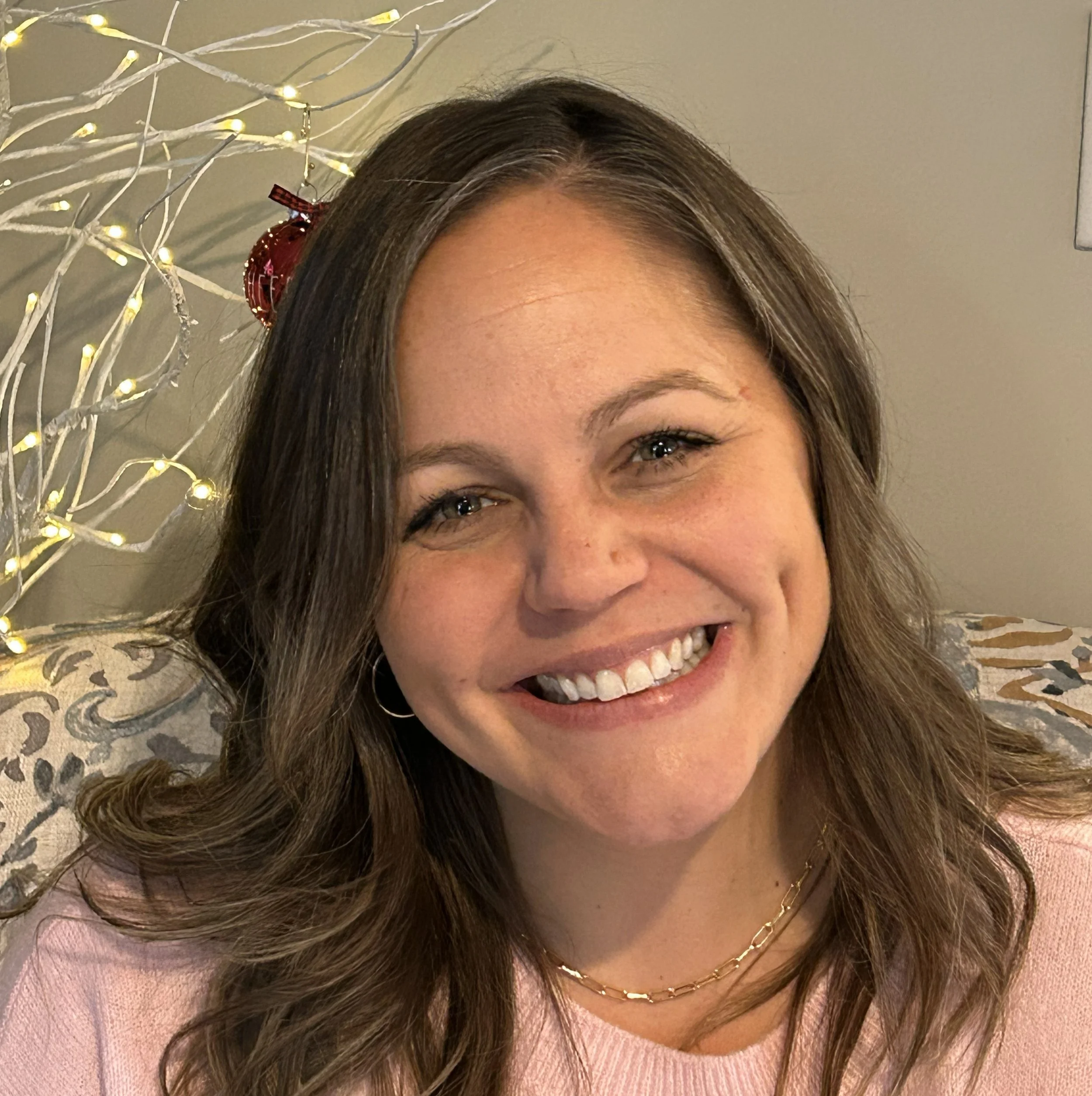 A smiling woman with wavy brown hair, wearing a pink top and a gold chain necklace, sitting in front of a decorated background with string lights and ornaments.