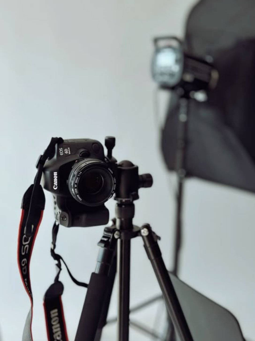 Camera mounted on a tripod, with a blurred bag and partial view of a light in the background against a plain wall.