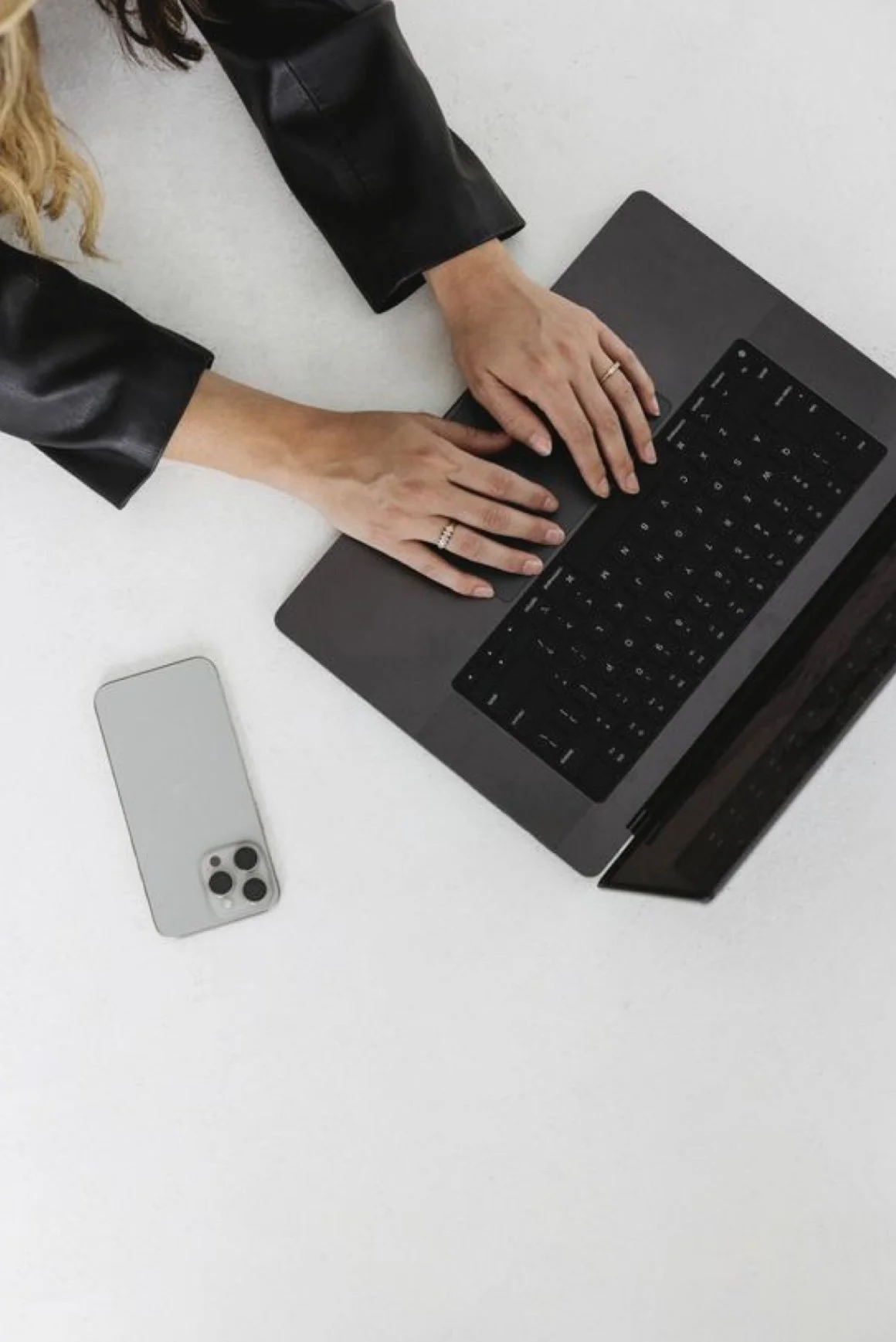 A person wearing a black leather jacket is typing on a laptop with a black keyboard. There is a silver smartphone placed on a white surface near the laptop.