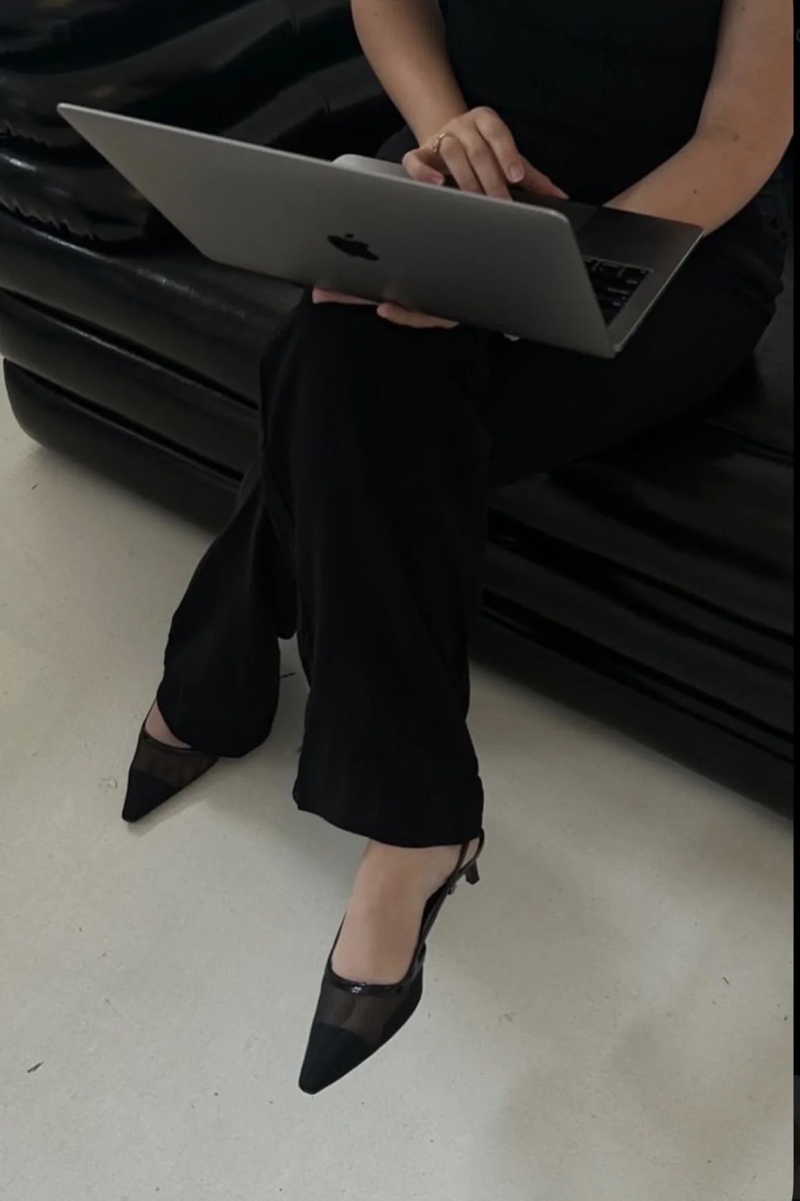 Person sitting on a black leather bench using a silver MacBook, wearing black pants, black pointed heels with sheer panels, and a black top.