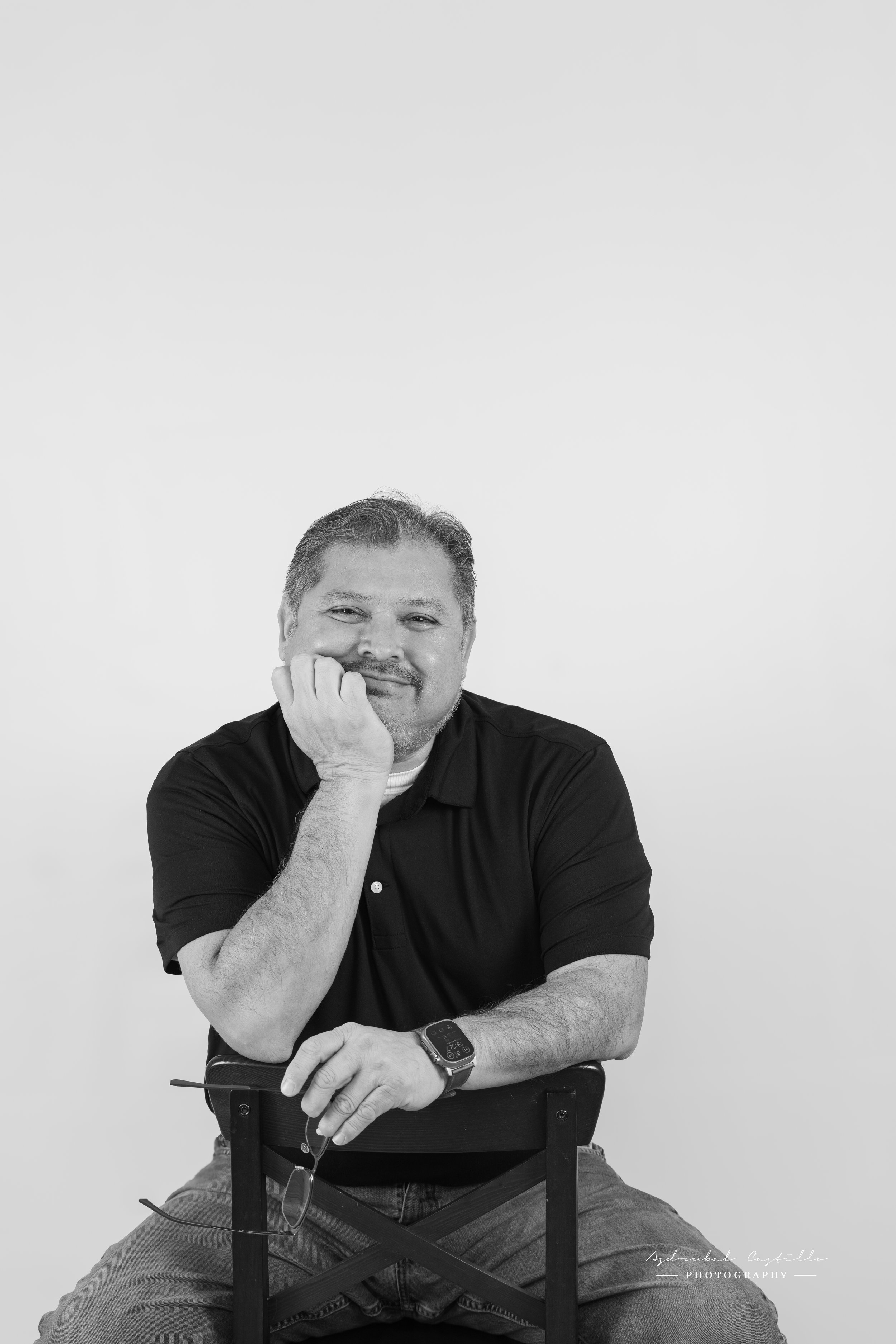 A smiling man with gray hair and beard, wearing a black polo shirt, sitting on a chair with his chin resting on his hand, holding glasses, with a smartwatch on his left wrist, against a plain white background.