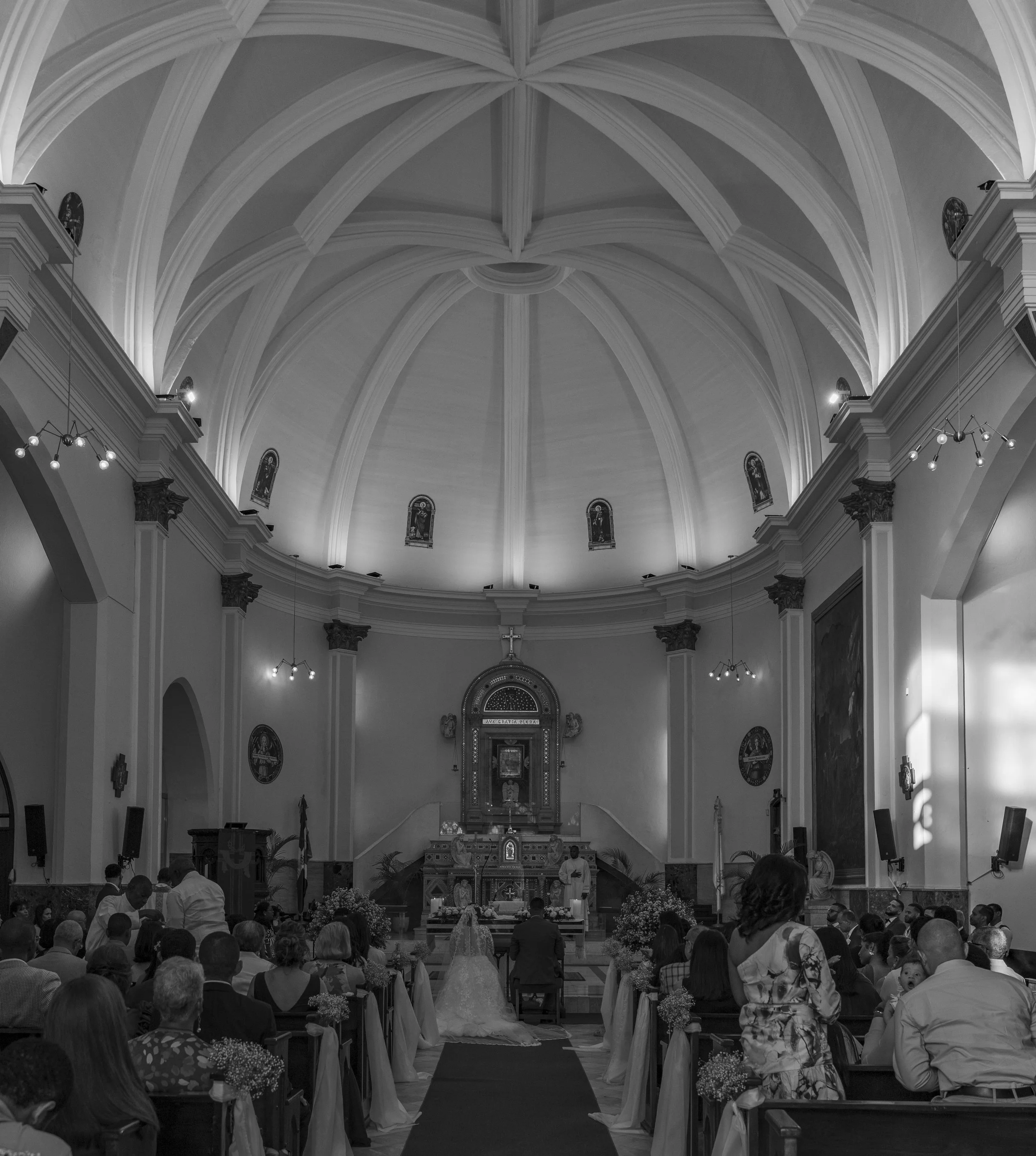 A black-and-white photo of a church interior during a wedding ceremony, with a bride and groom at the altar and guests seated on both sides.