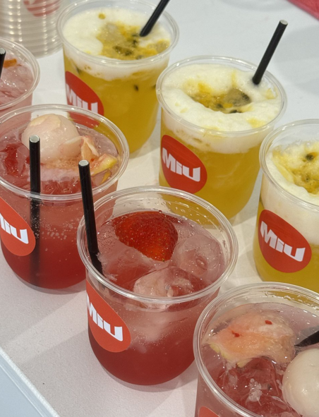 Several cups of colorful fruit-flavored beverages with ice, some topped with fruit pieces, on a white table.