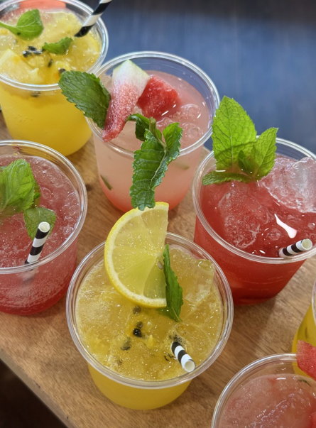 Six colorful fruit drinks with ice, garnished with mint leaves, fruit slices, and striped straws on a wooden surface.