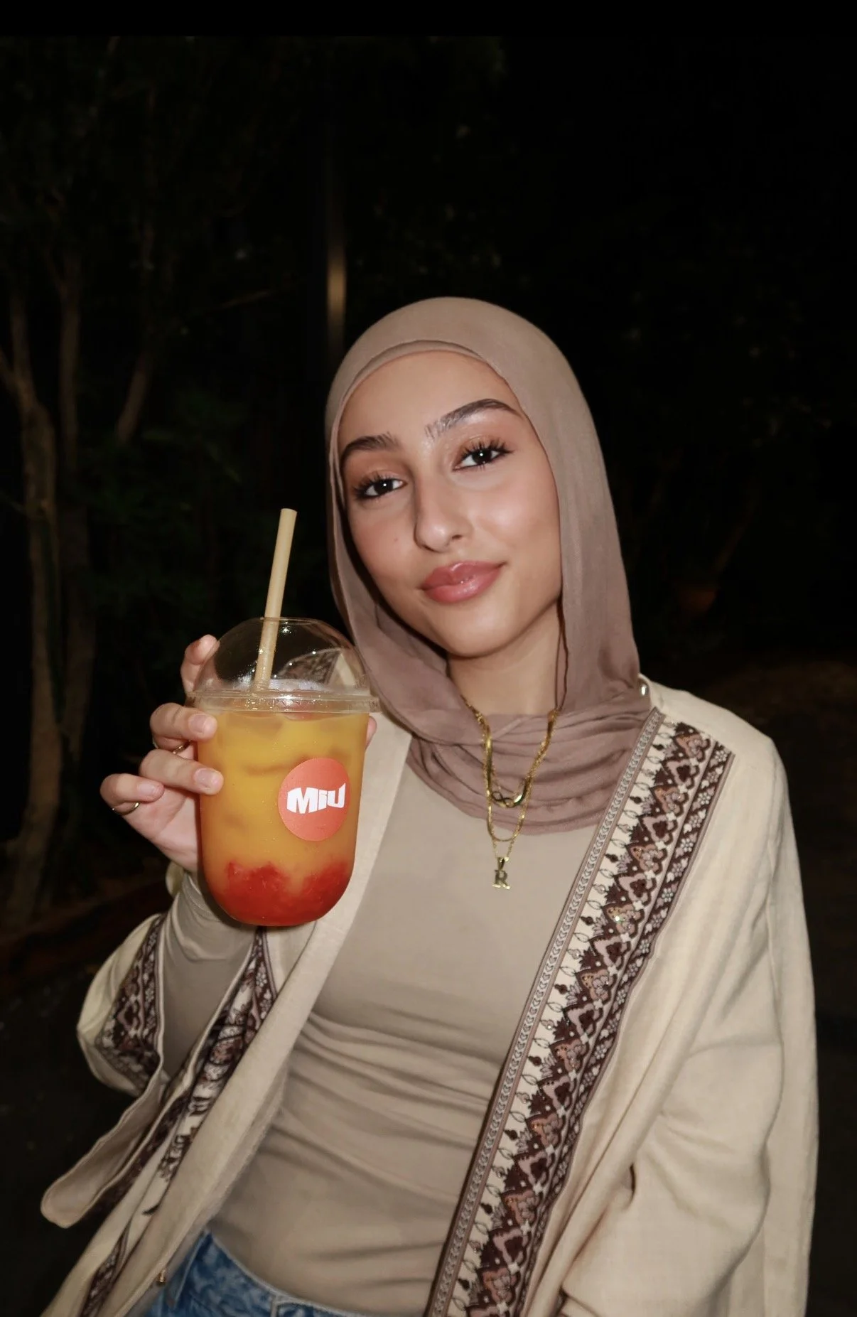 A young woman wearing a beige hijab and beige clothing holding a colorful beverage with ice and fruit slices, smiling at the camera at night outdoors.