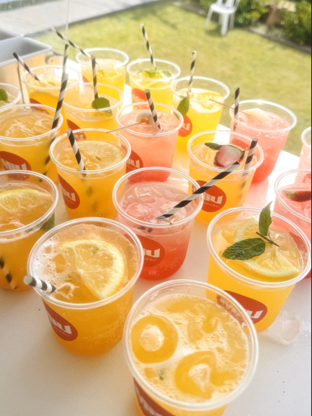 Multiple cups of colorful fruit-infused beverages with lemon, mint, and strawberries, some with black and white striped straws, arranged on a white table outdoors.