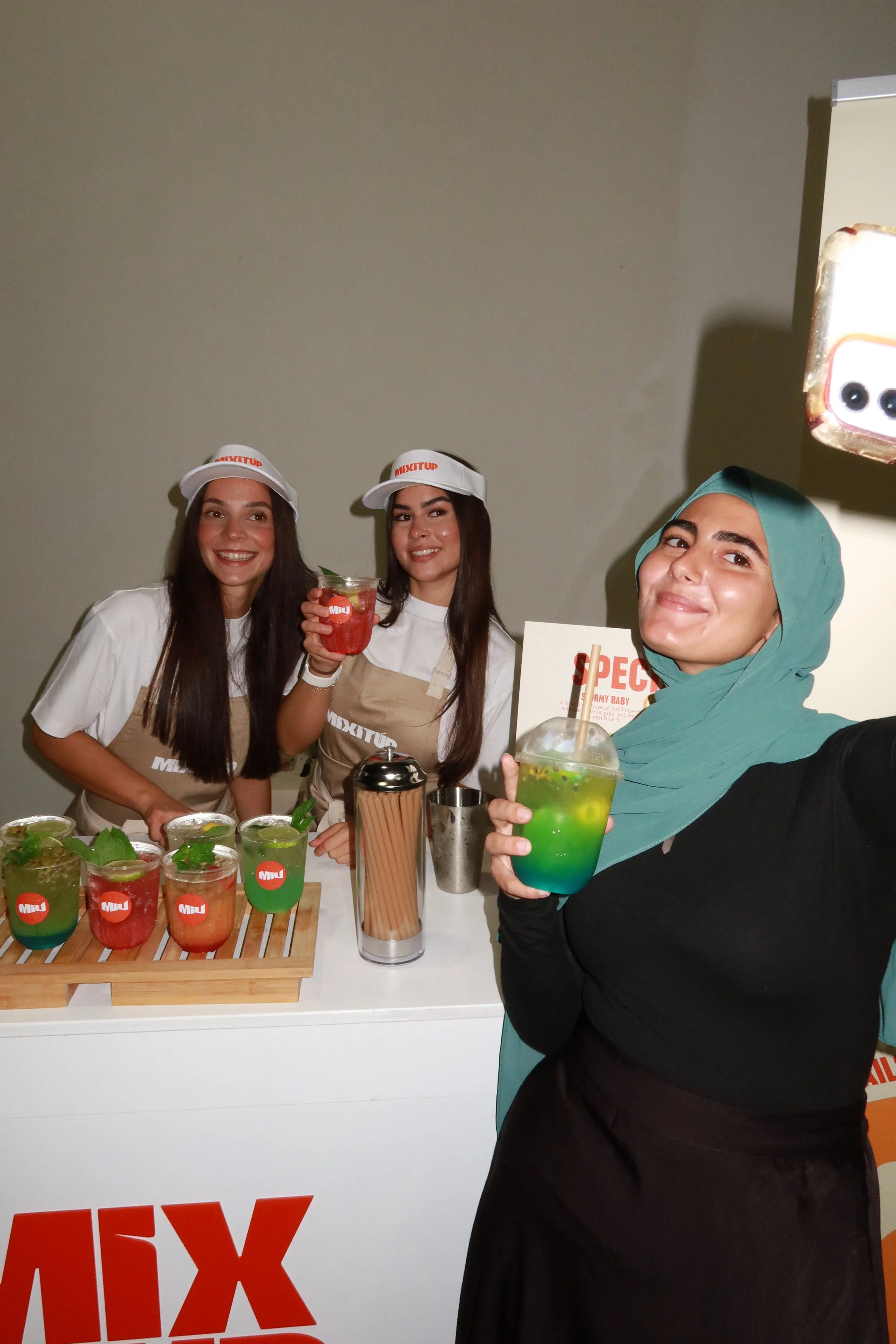 Three women behind a drink stand selling colorful beverages, two women wearing white caps and aprons, and one woman with a teal headscarf holding a layered drink, smiling at the camera.