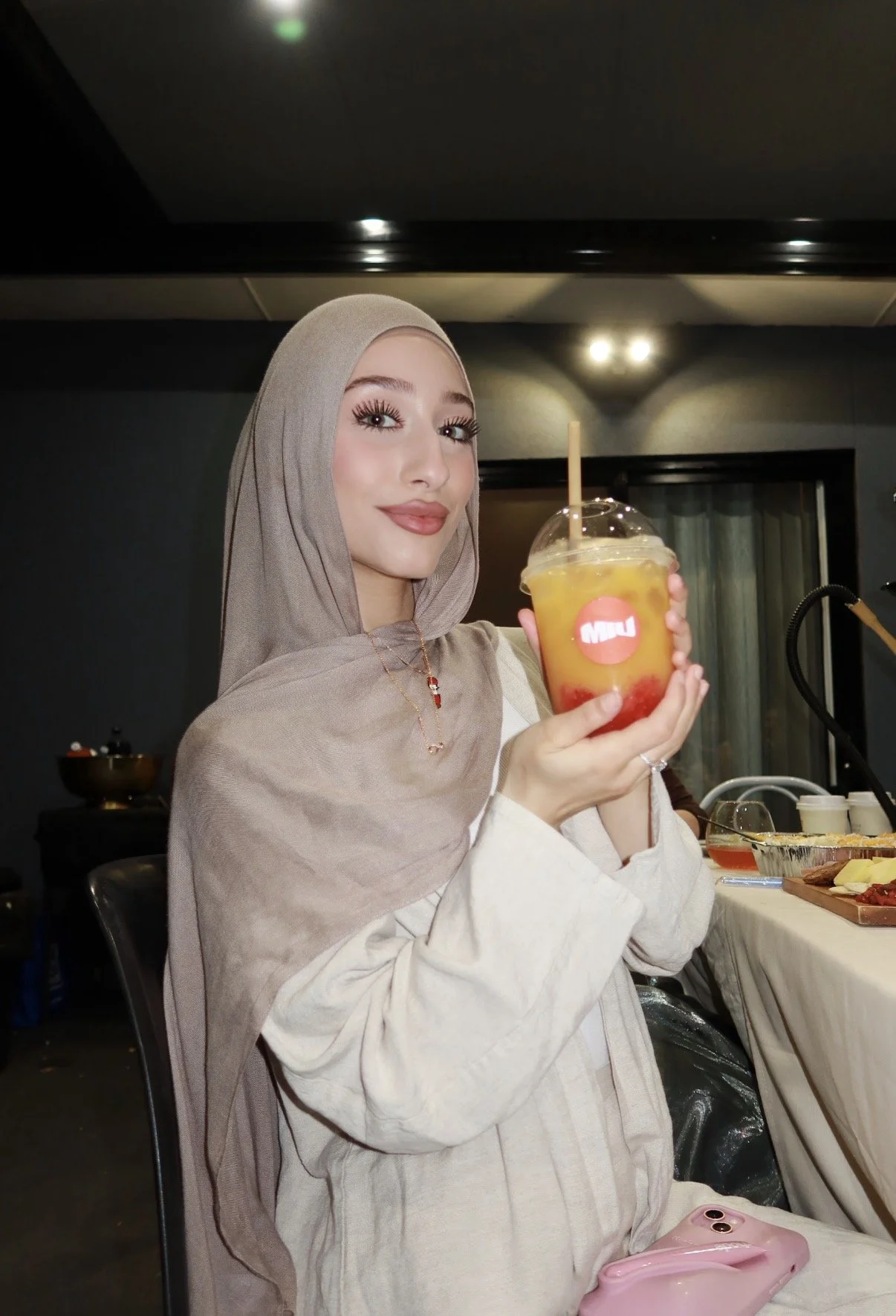 A woman wearing a beige hijab and cream-colored outfit holding a large cup of yellow and red fruit drink with a straw.