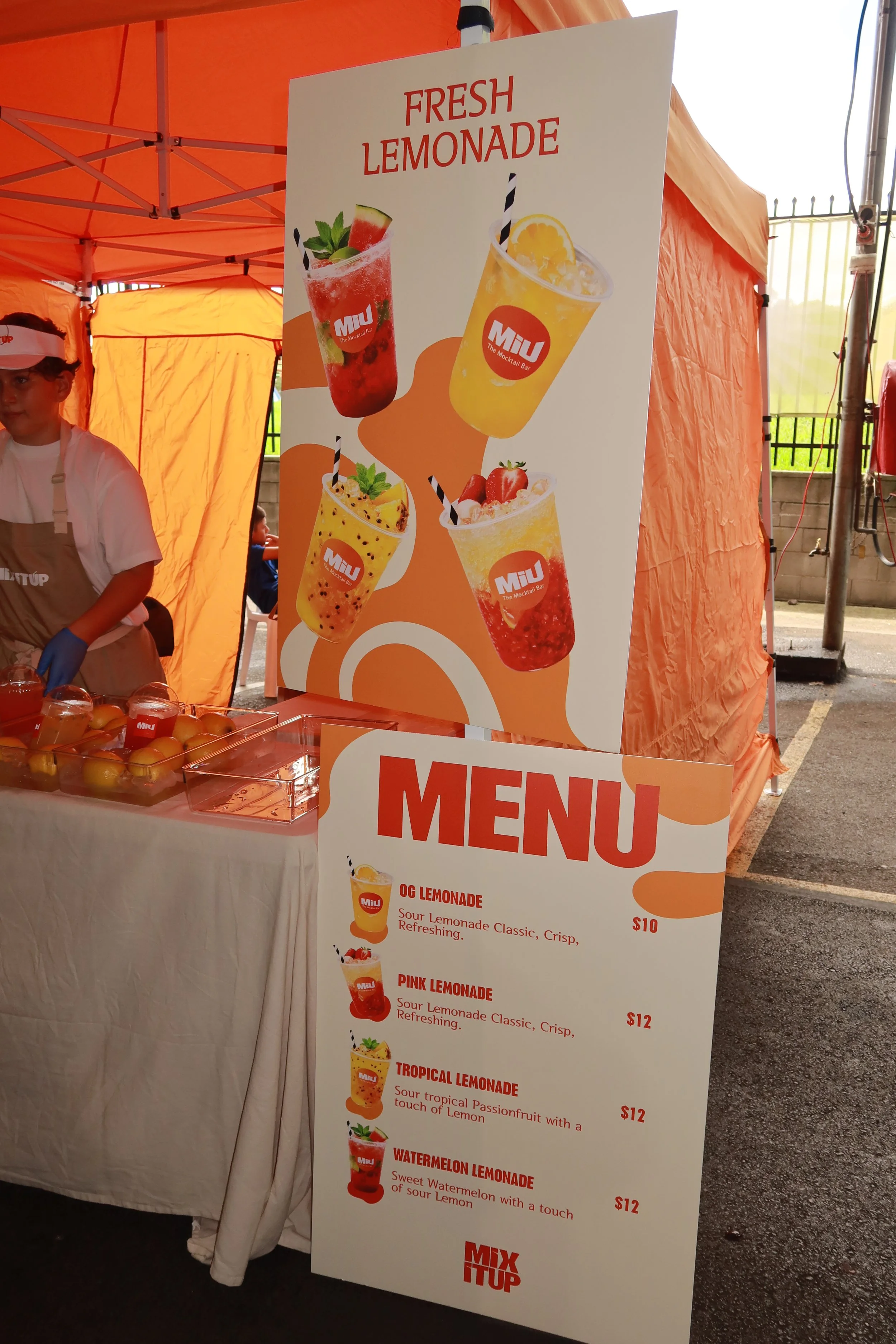 A large sign advertising fresh lemonade with pictures of four different lemonade drinks, inside a stand with an orange canopy. The menu below lists four flavors: OG Lemonade, Pink Lemonade, Tropical Lemonade, and Watermelon Lemonade, with prices rang