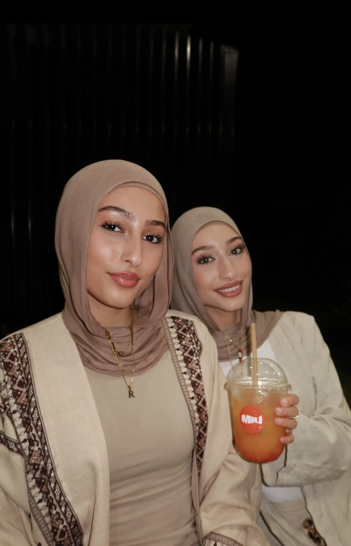 Two women wearing beige hijabs and traditional beige clothing smiling at the camera; one woman is holding a beverage in a clear cup with a straw.