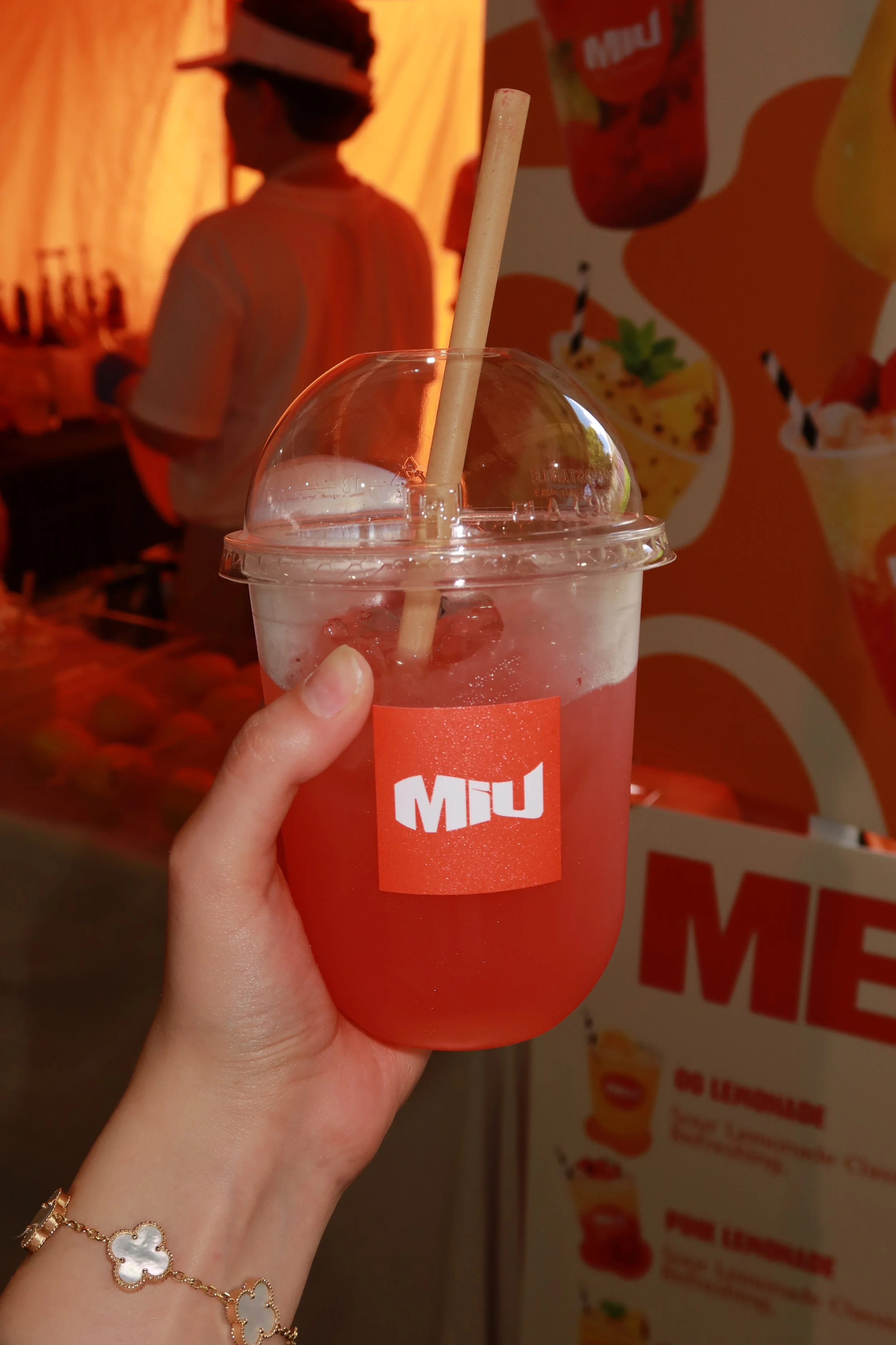 A hand holding a clear plastic cup filled with a pink-colored beverage and ice, with a straw, in front of a bright, colorful background featuring a man in a hat, a promotional poster for a dessert shop, and images of drinks and desserts.