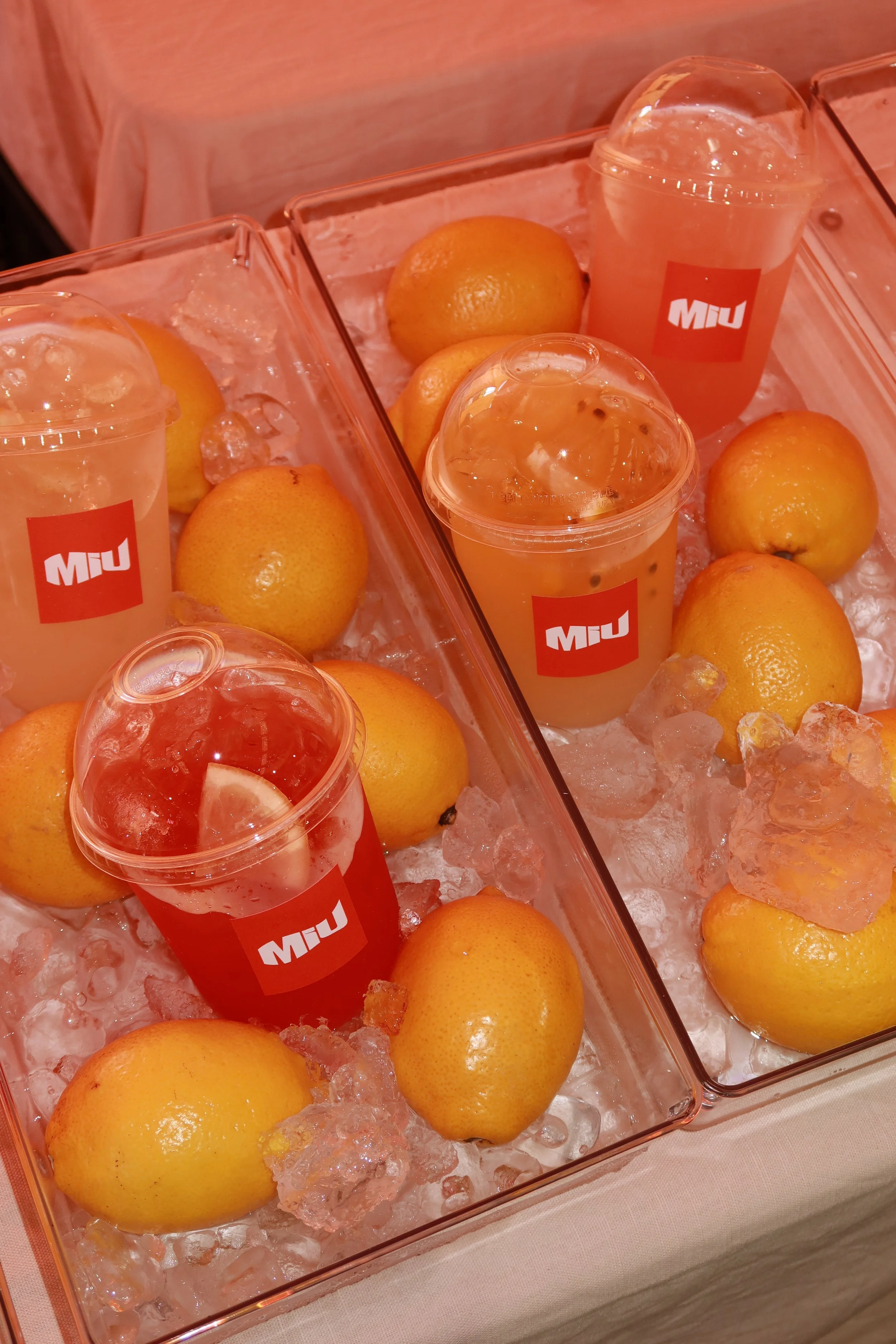 Six clear plastic cups with red MIU branding, filled with pink and yellow beverages, placed on ice alongside fresh lemons in glass containers.