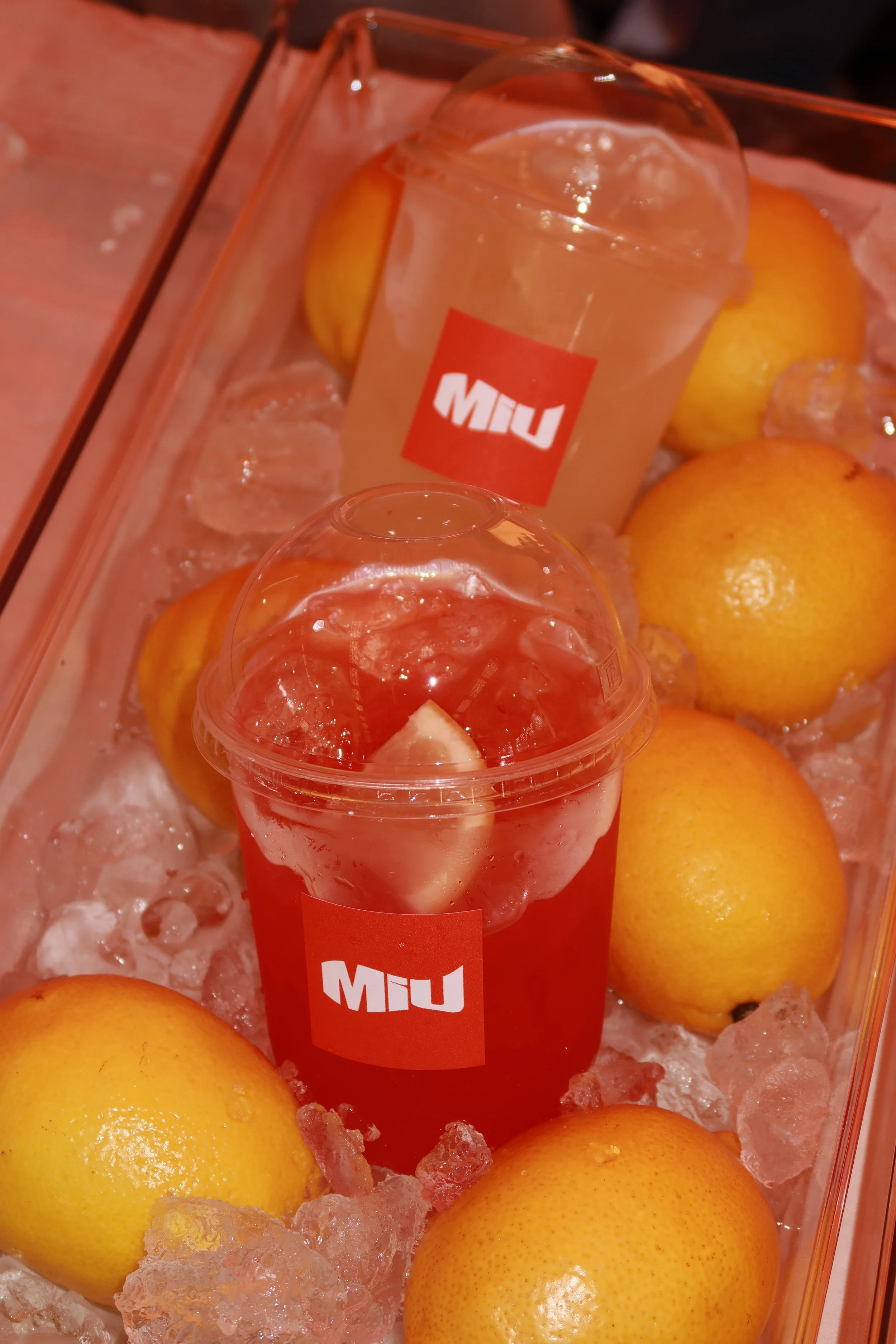 Two transparent cups of lemonade with ice and lemon wedges, placed on a bed of ice alongside whole lemons inside a glass container.