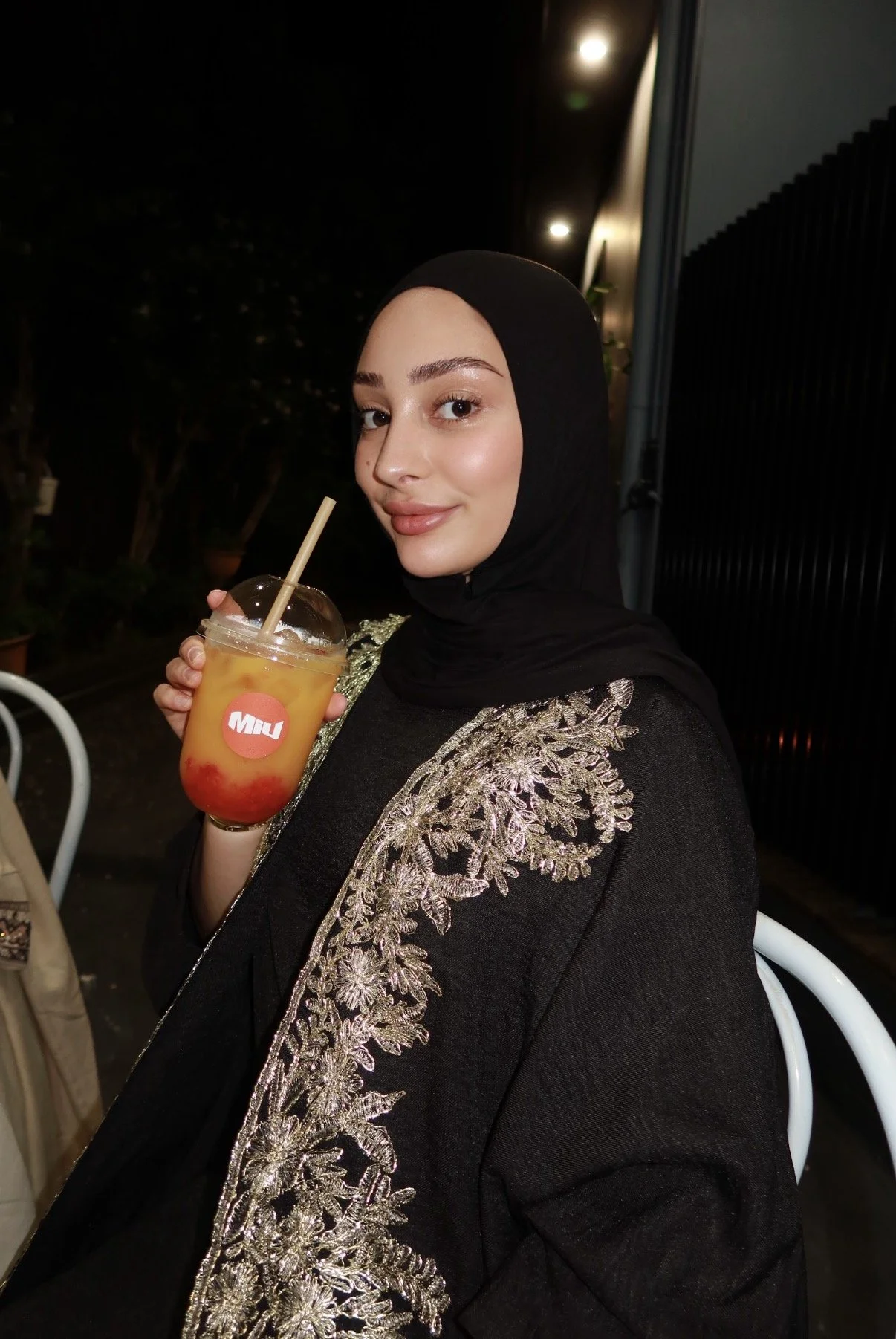 A woman wearing a black hijab and a black garment with gold embroidery is holding a colorful drink with a straw. She is smiling at the camera in an outdoor setting at night.