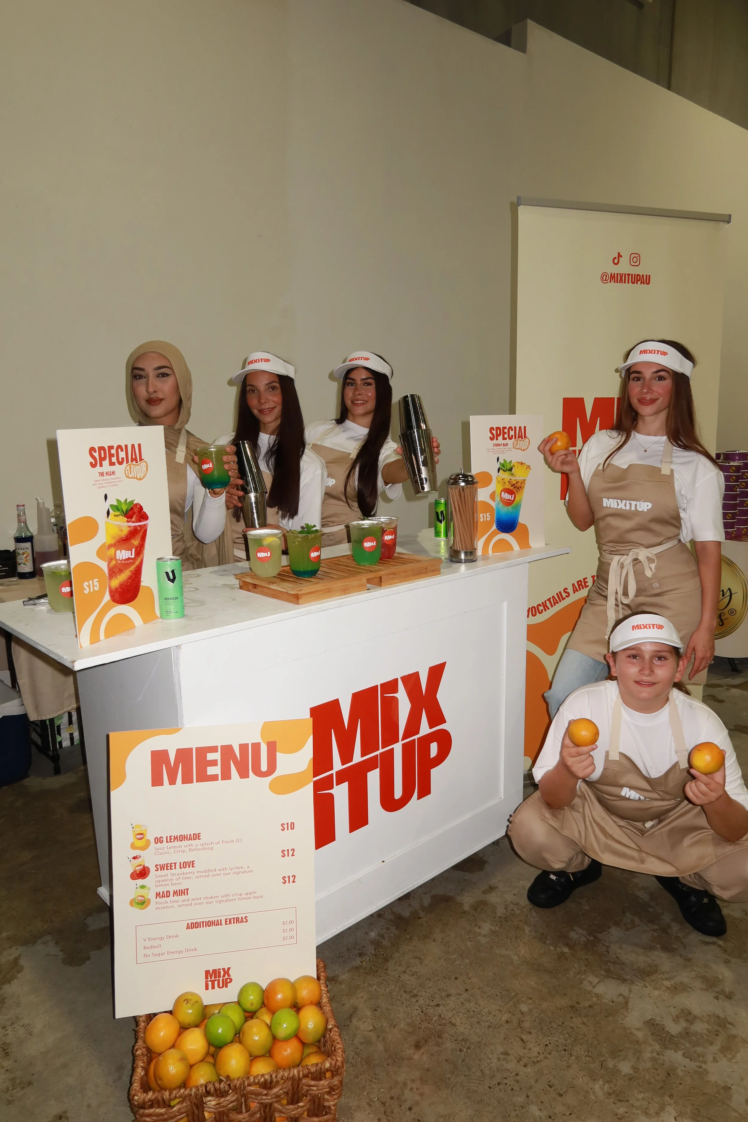 Group of five young women at a juice stand called 'Mix It Up,' wearing aprons and hats with the brand logo, surrounded by colorful drinks, fruits, and a menu board with prices.