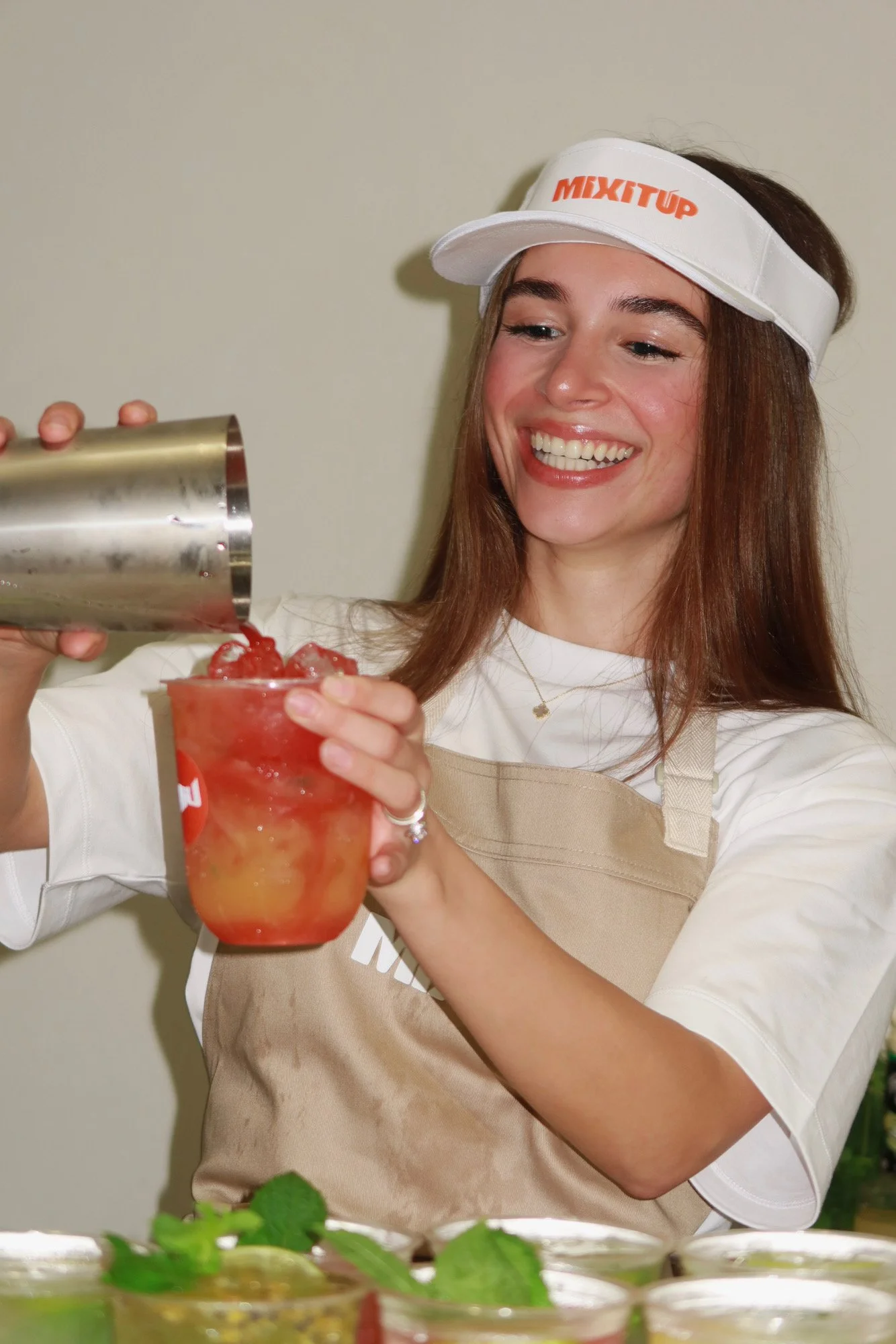 A woman smiling and pouring a red drink into a glass, wearing a white visor with 'MIXITUP' written on it and a beige apron.