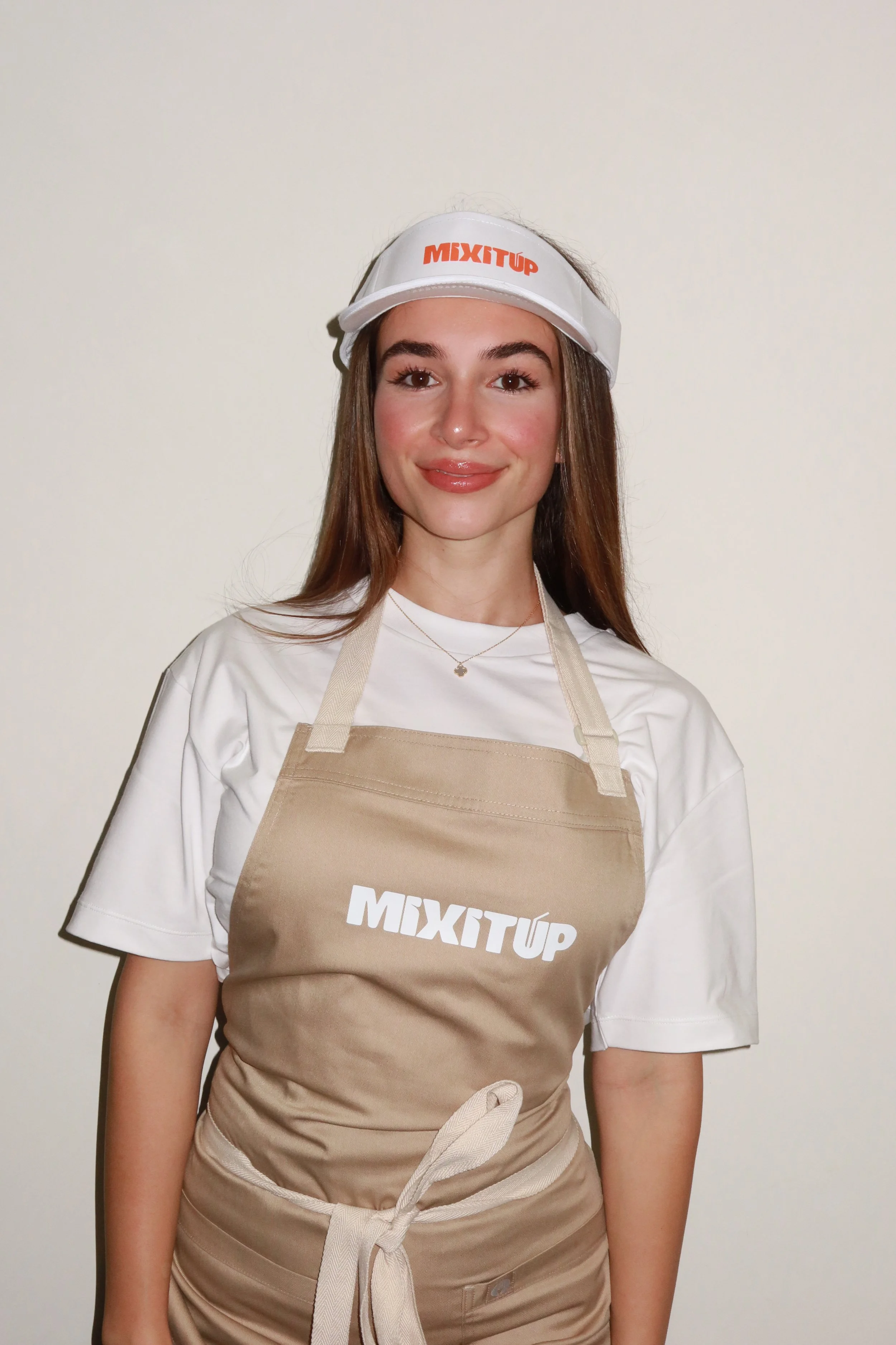 A smiling woman wearing a white visor and T-shirt, beige apron with 'MIXTUP' logo, standing against a plain white wall.