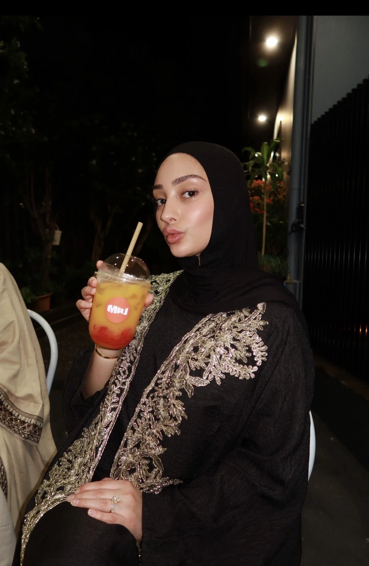 A woman wearing a black hijab and black dress with gold embroidery is sitting outdoors at night, holding a colorful drink with red and yellow layers, a straw, and a red sticker with white text.