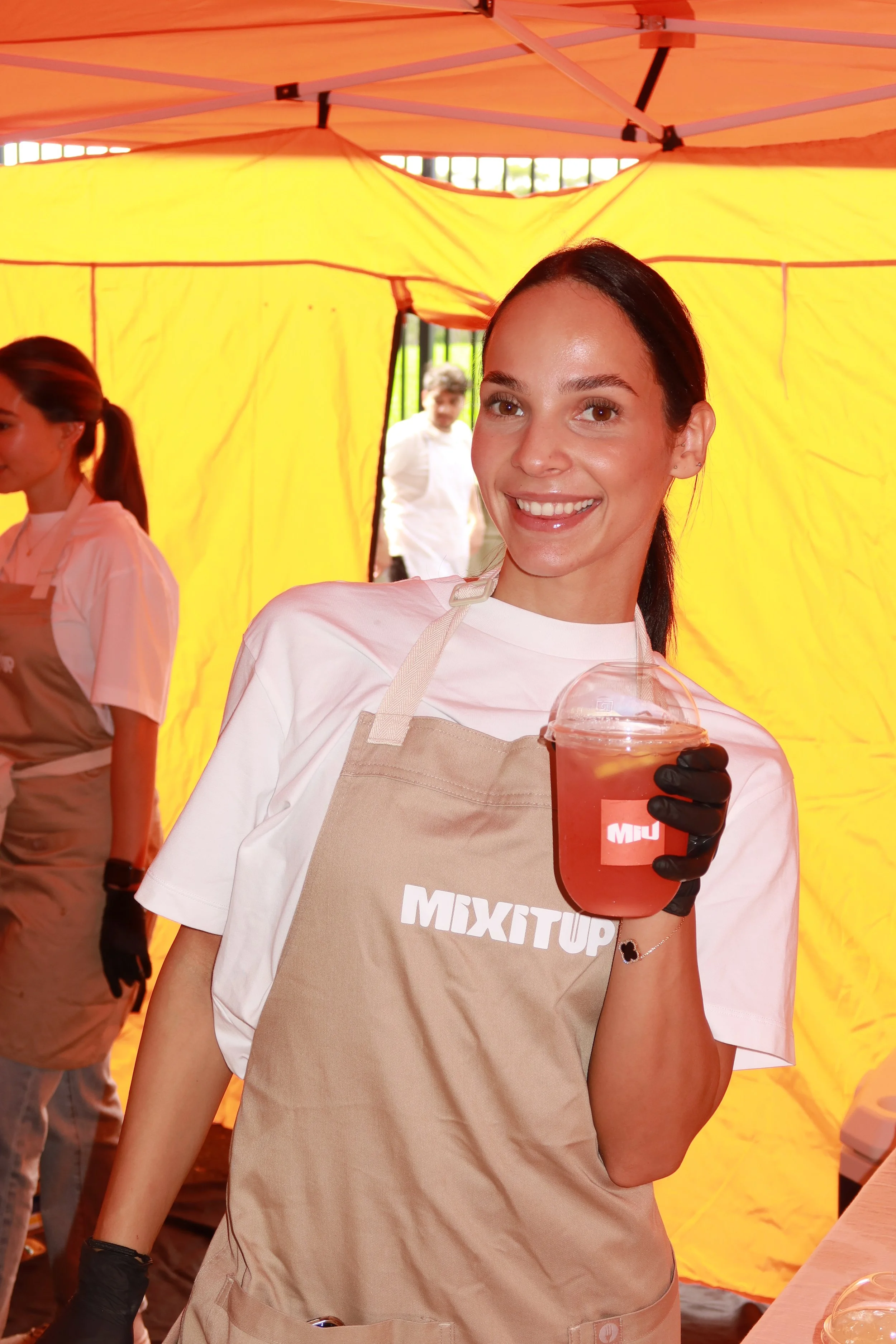A woman smiling and holding a pink beverage in a clear plastic cup, wearing a beige apron with the word 'MIXTUP' on it, at an outdoor event with a yellow tent in the background.