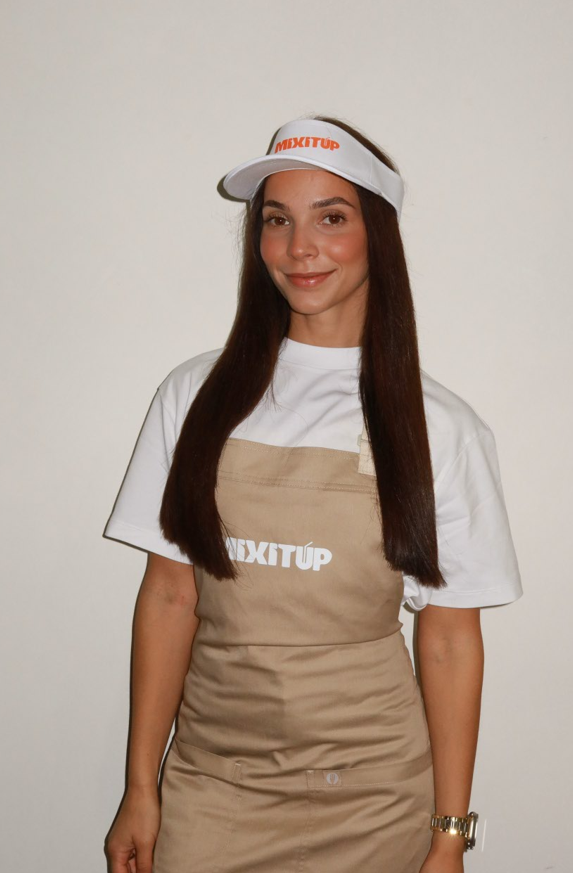 A young woman wearing a white visor with 'MIXITUP' written on it, a white t-shirt, and a beige apron with 'MIXITUP' on it, standing against a plain white background.