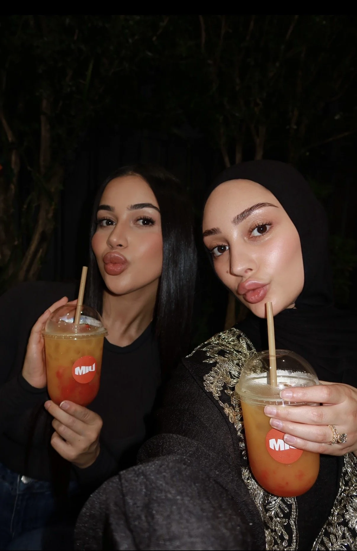 Two women taking a selfie and holding colorful fruit drinks with straws, one wearing black and the other wearing a black headscarf with gold embroidery, outdoors at night.