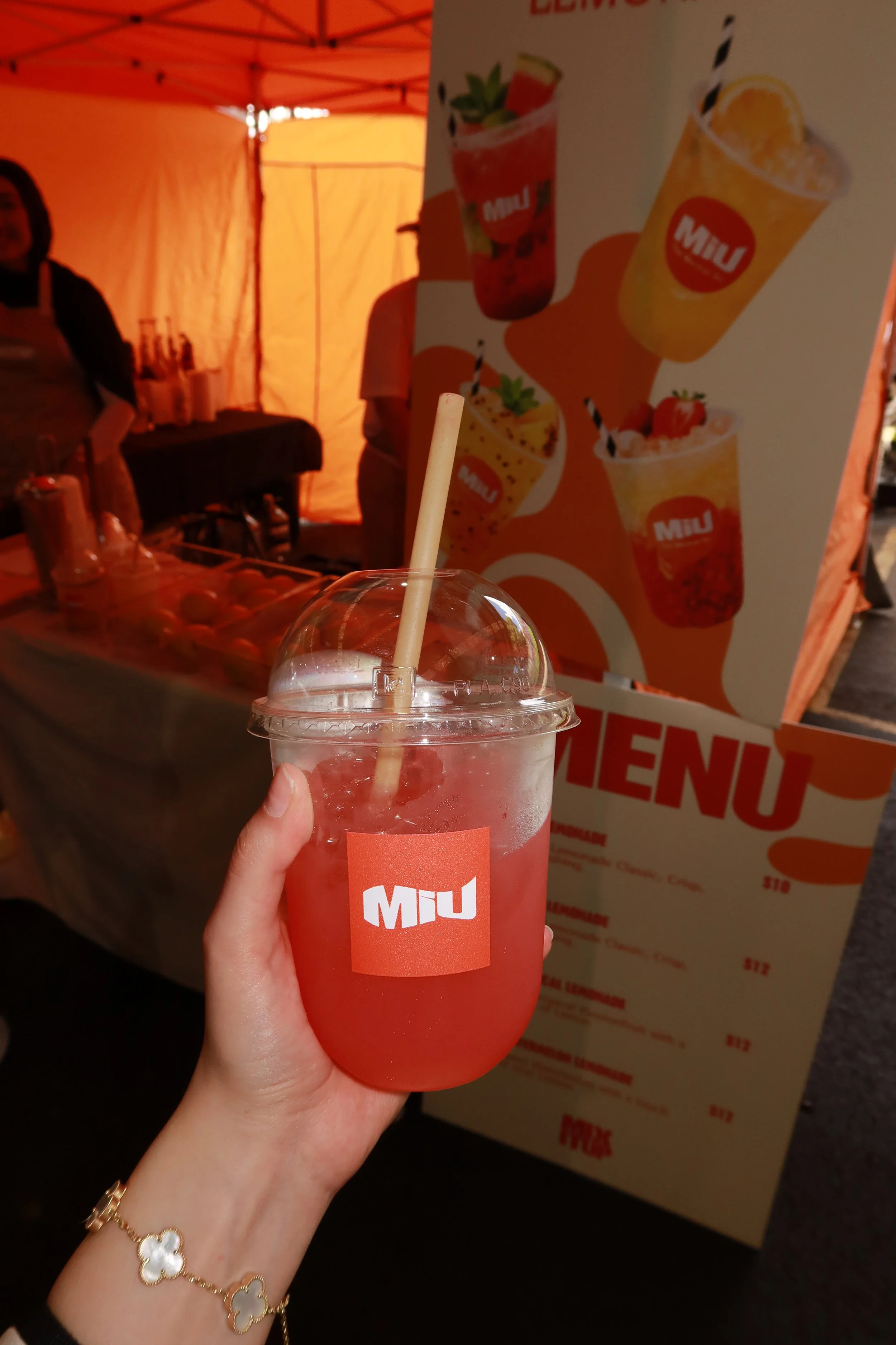 Person holding a pink fruit drink in a clear plastic cup with a straw, branded with a red logo reading 'MIU', at an outdoor food stall with a menu and a large drink advertisement in the background.