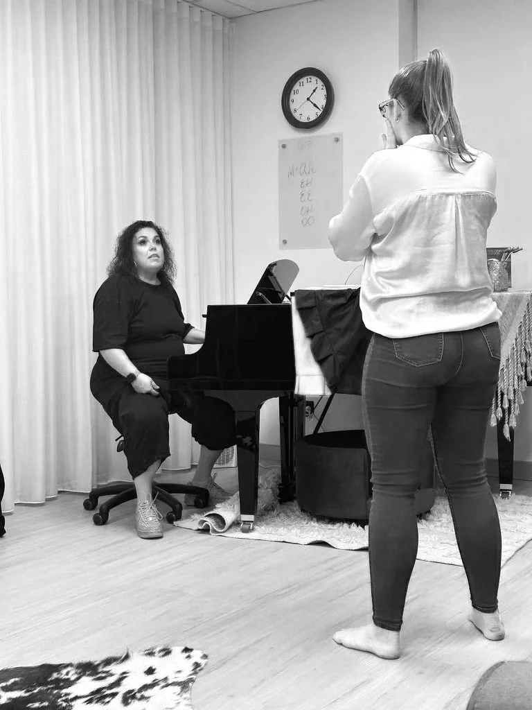 Vocal Coach Mindy Pack teaching Emma McGuire. Mindy is sitting at a piano with sheet music, Emma is standing and facing her.