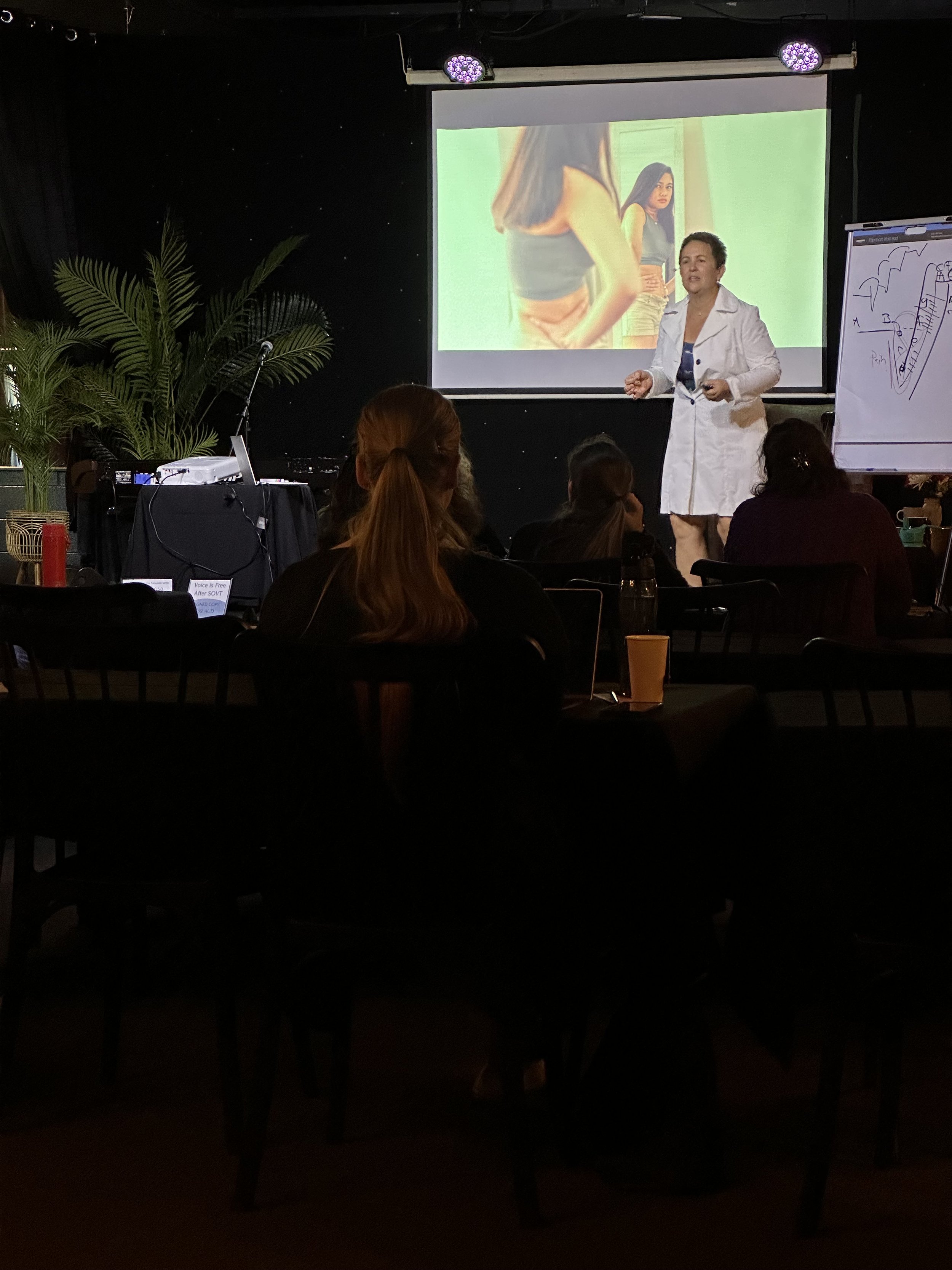 A woman in a white coat giving a presentation in front of a projected image of two dark-haired women in casual clothing. Audience members are seated at tables in a dimly-lit room with plants and a flip chart to the side.