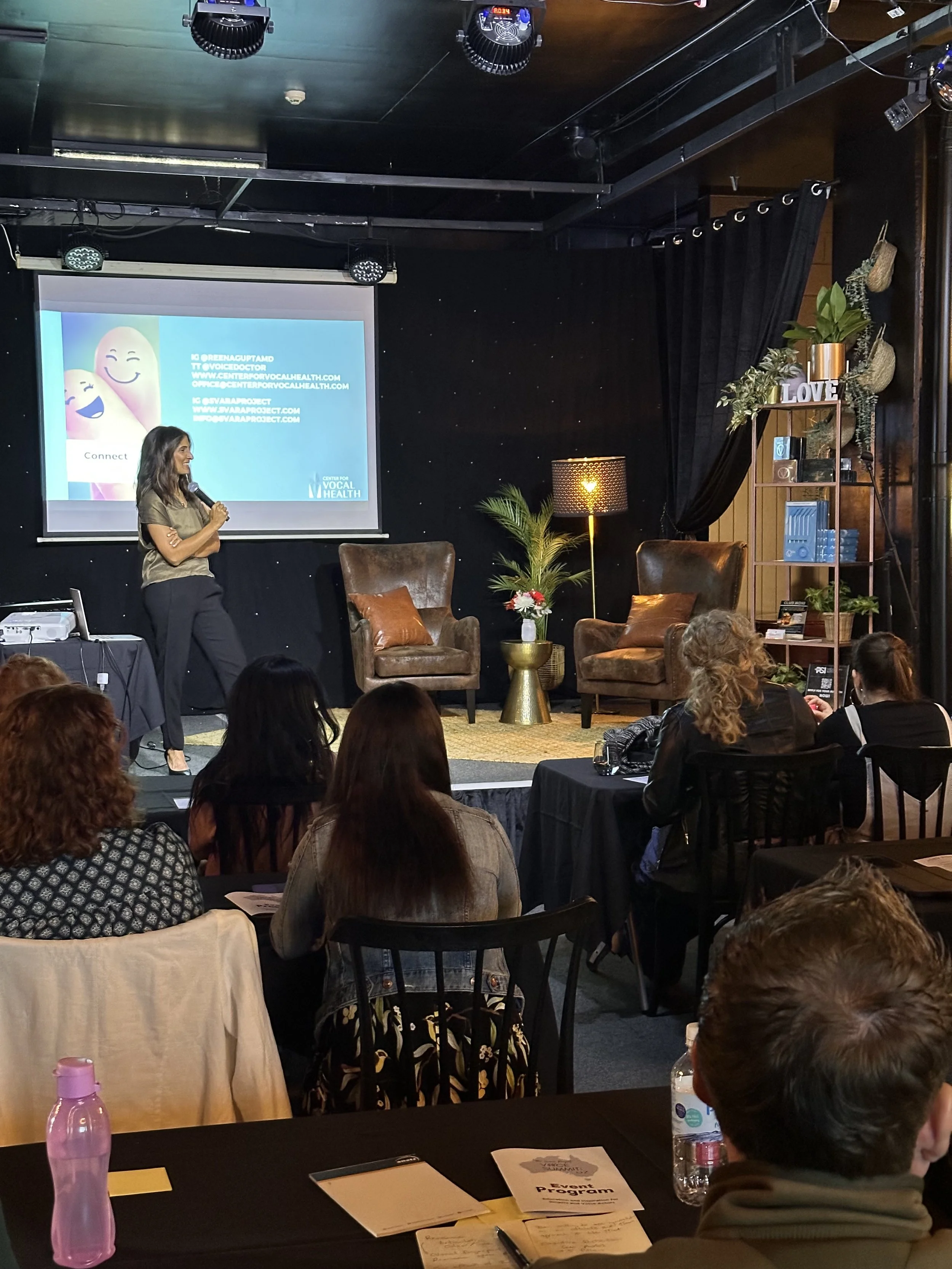 A woman stands on a stage giving a presentation in front of an audience in a dimly lit room. She is holding a microphone and smiling. The background features a large screen with a slide, two leather armchairs, a floor lamp, and decorative shelves with plants and decor.