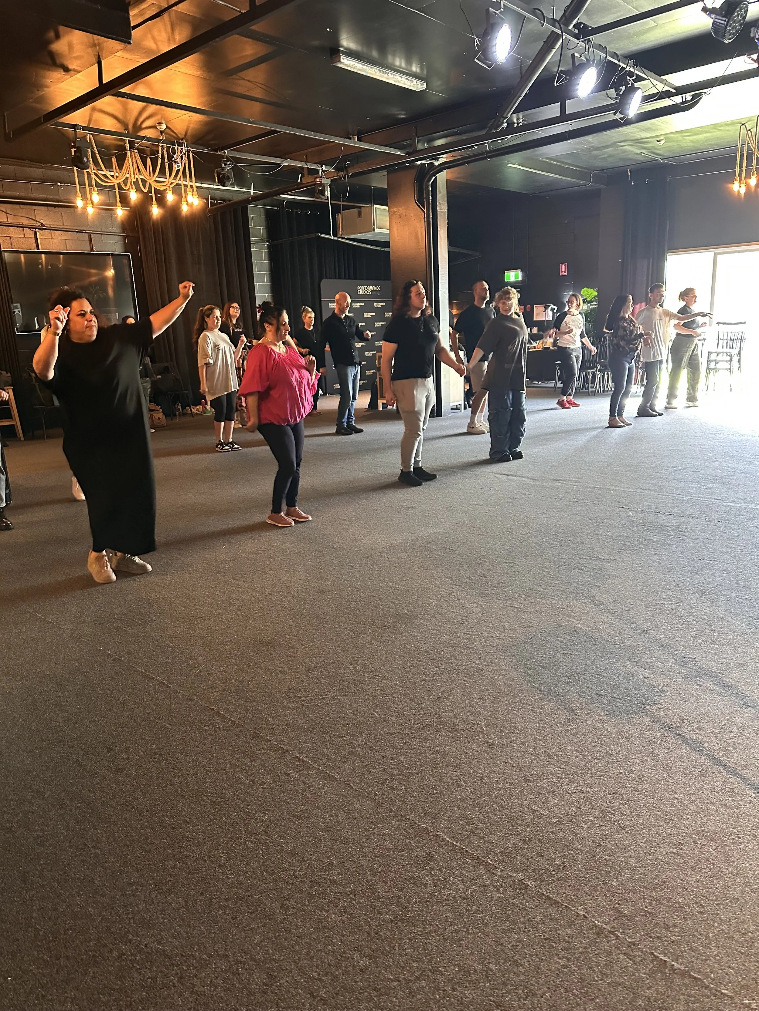 Group of people practicing line dancing in a spacious indoor area with black walls and large windows, illuminated by overhead lights.