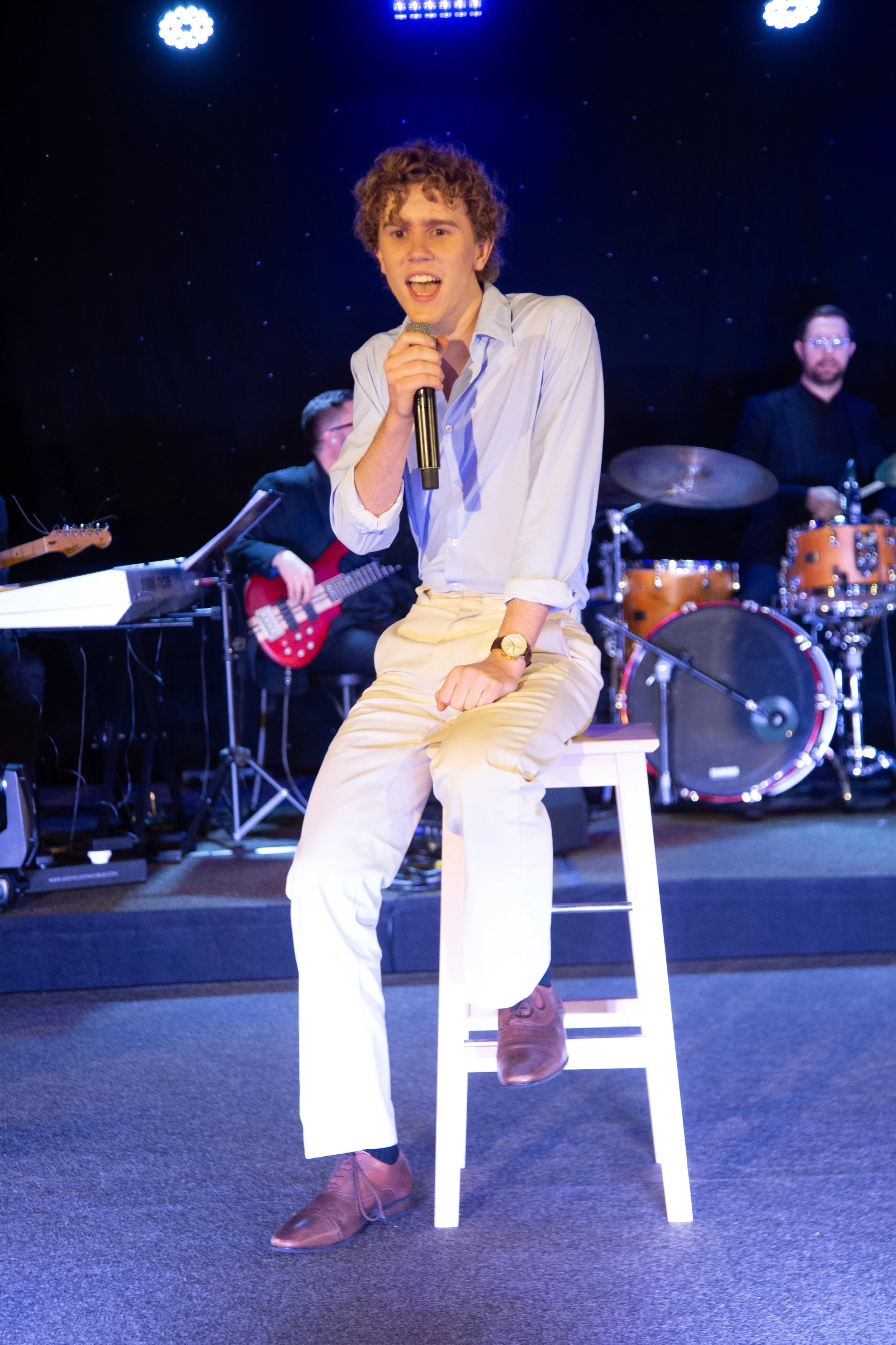 A young man singing into a microphone on stage with a band in the background, including a guitarist, keyboardist, and drummer, during a live performance.