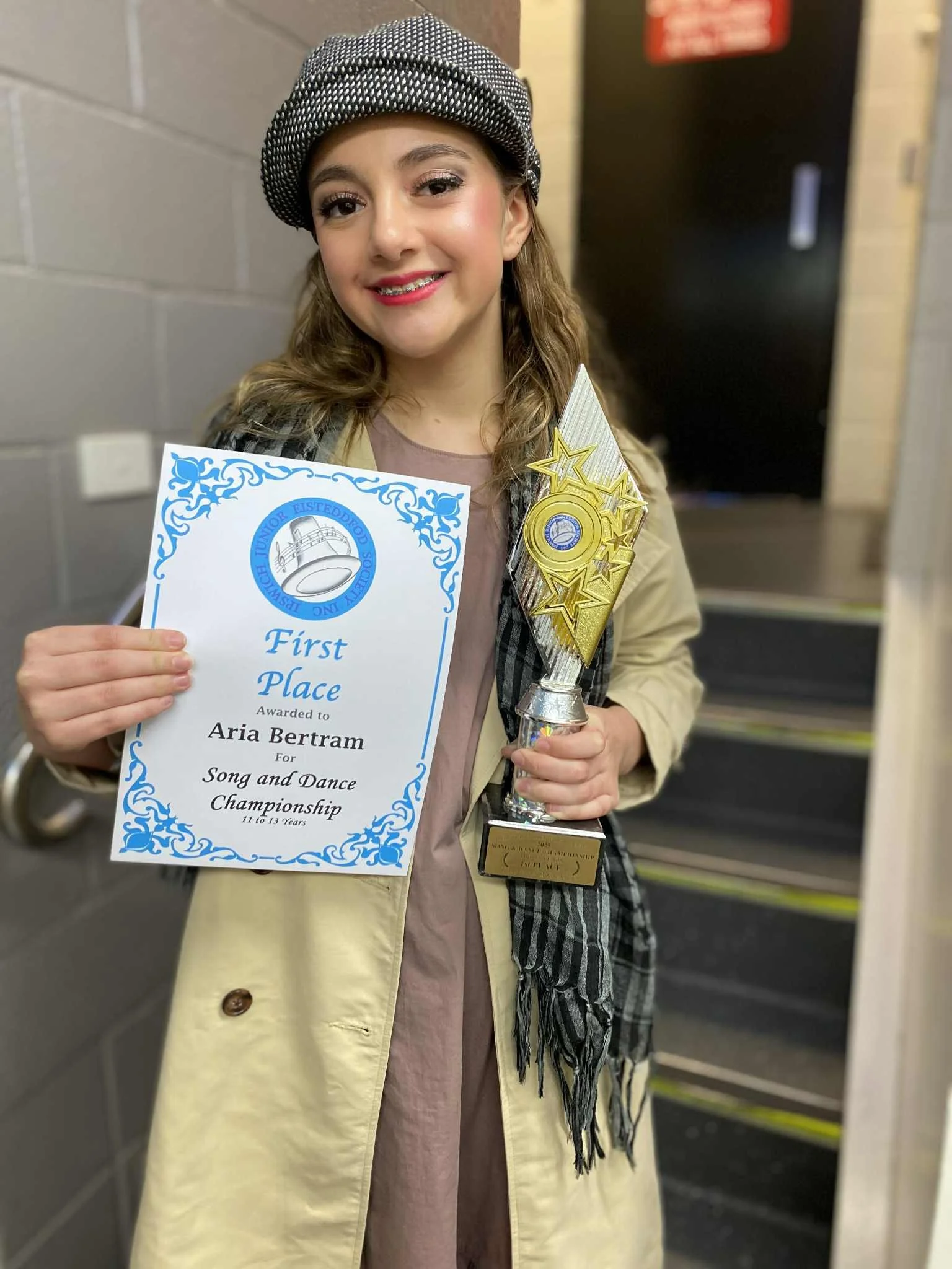 A young girl with wavy brown hair, wearing a grey beret hat, beige trench coat, and pink lipstick, holding a trophy and a certificate. The certificate reads 'First Place' awarded to Aria Bertram for Song and Dance Championship, ages 11-13. She is smiling and standing on a staircase indoors.