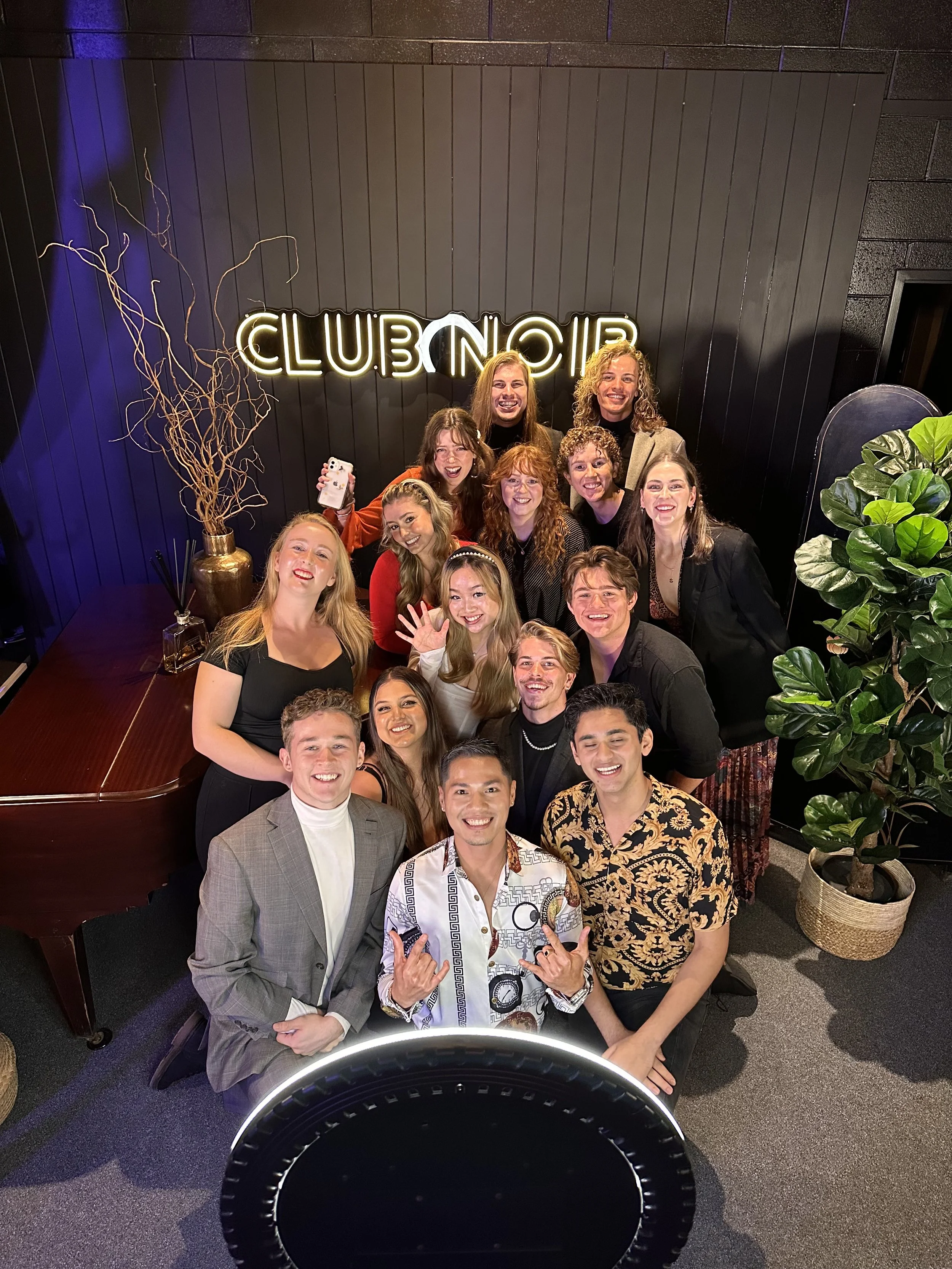 A group of young adults smiling and posing for a photo at a club or lounge, in front of a neon sign that reads 'CLUB NOIR' for the Rise Up Senior Semi Finals in 2024.