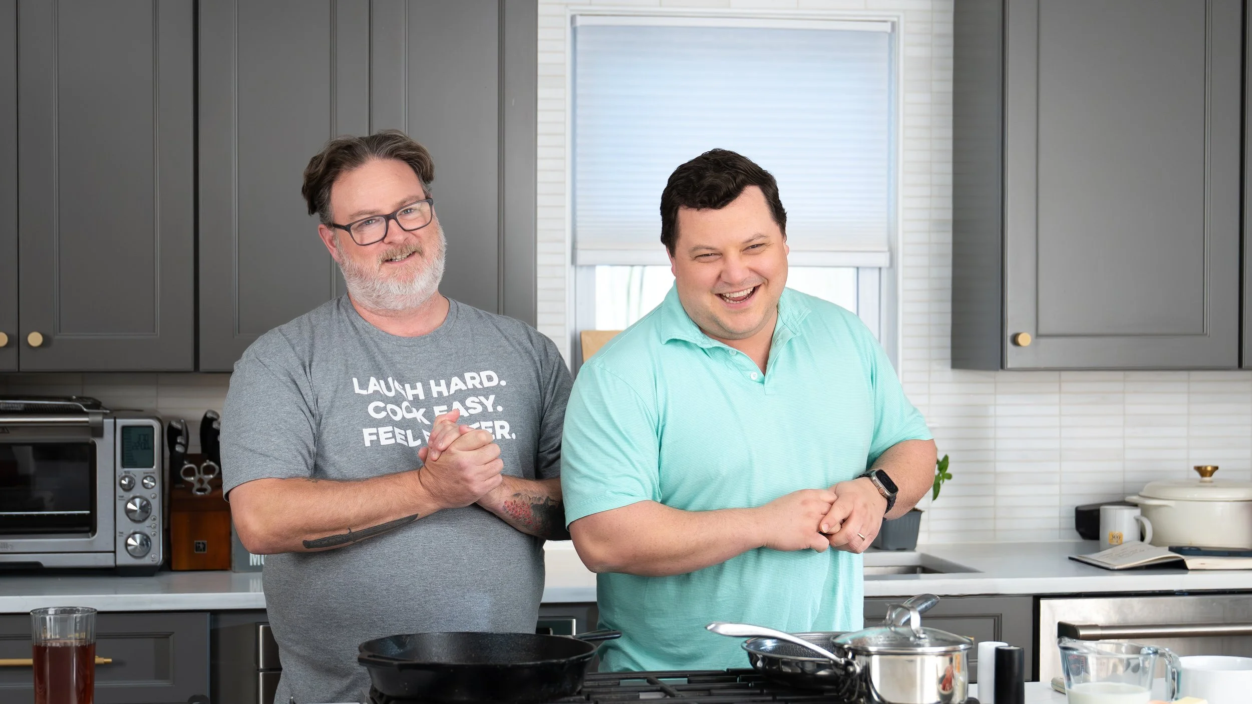 Two men stand in a kitchen, one with glasses and a beard clapping, the other smiling with folded hands. There are kitchen appliances and utensils on the counter.