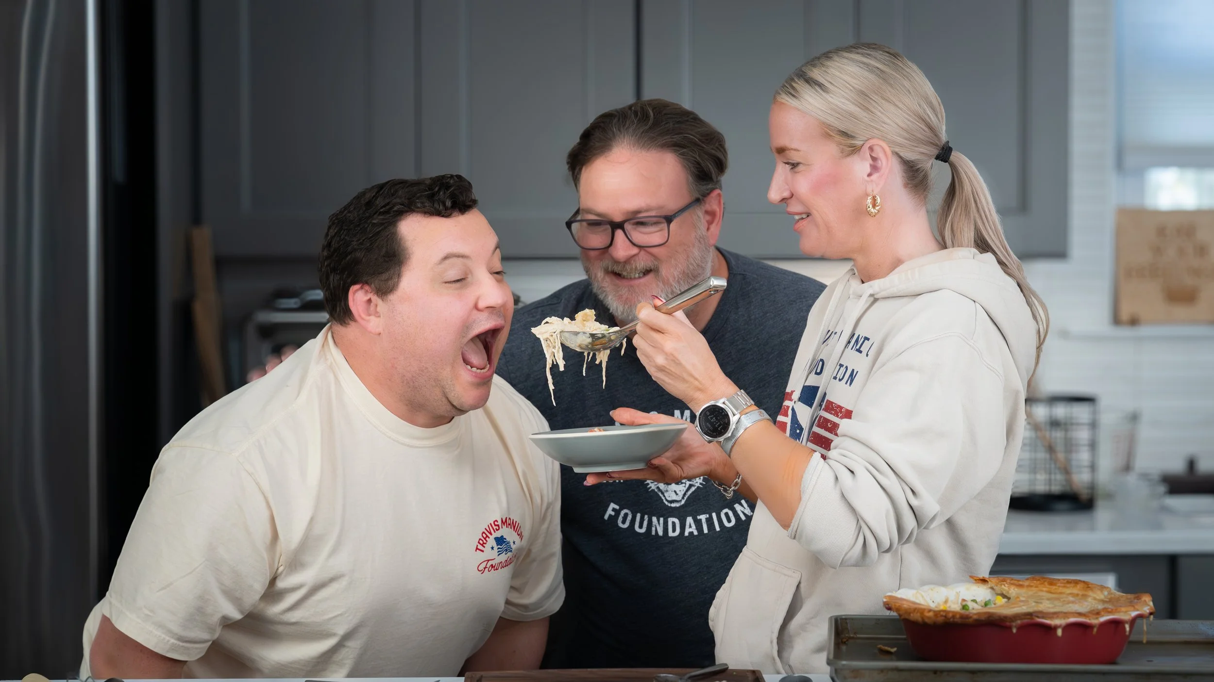 Three people sharing a meal in a kitchen, one woman feeding a man with a spoon, all smiling and laughing.
