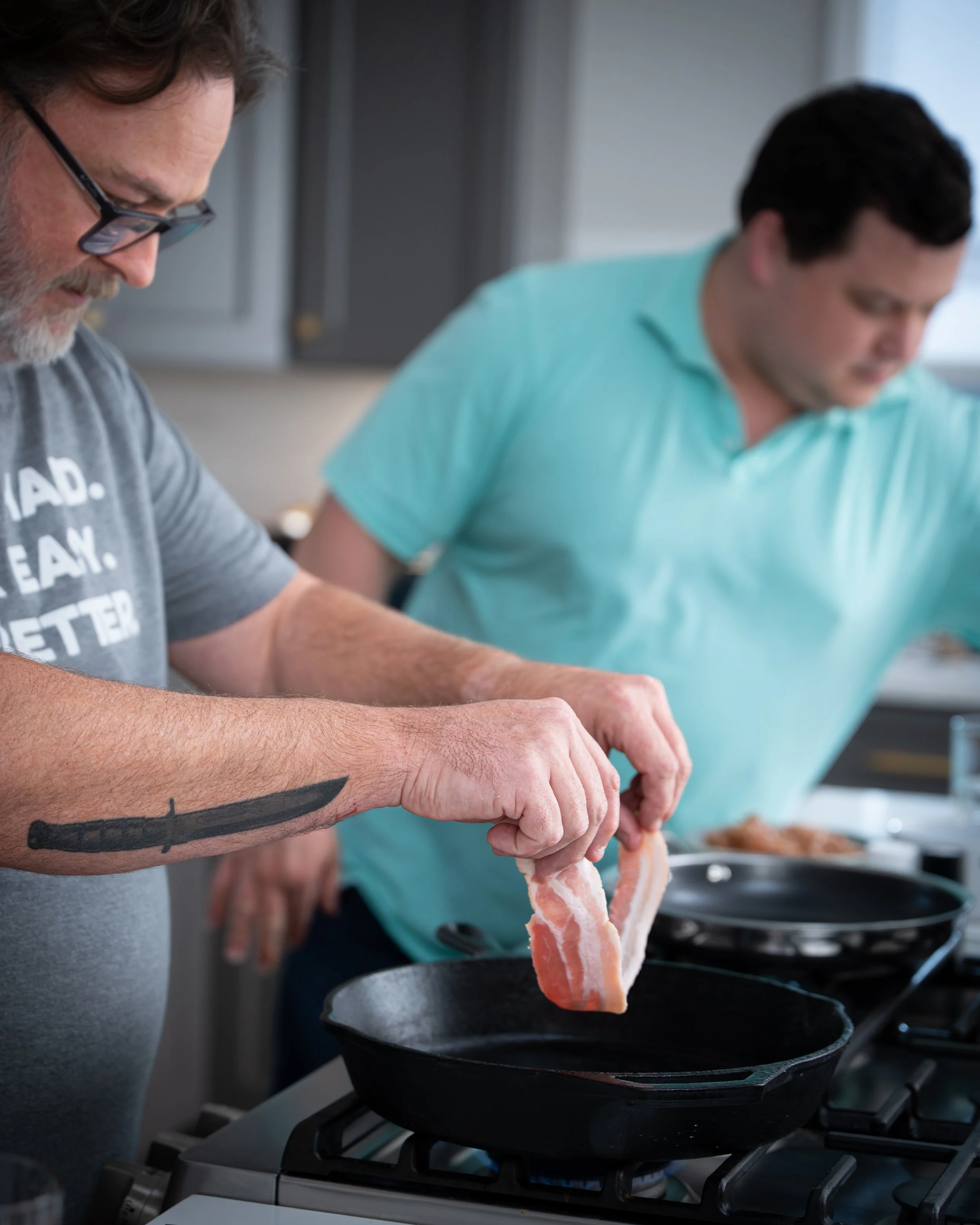 Two men cooking in a kitchen, one placing bacon in a frying pan on a stove.