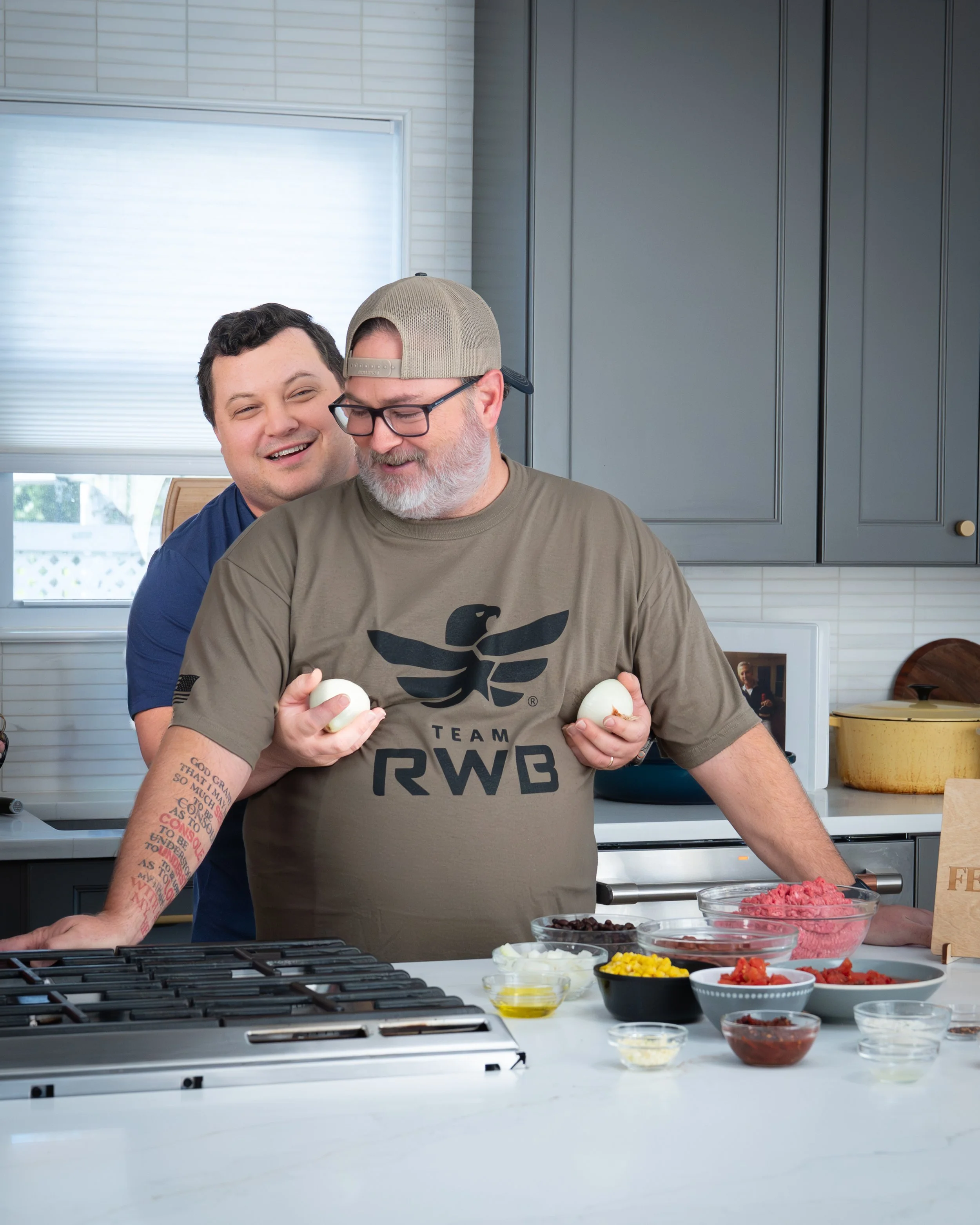 Two men in a kitchen, one holding eggs and the other smiling, with various ingredients on the counter.