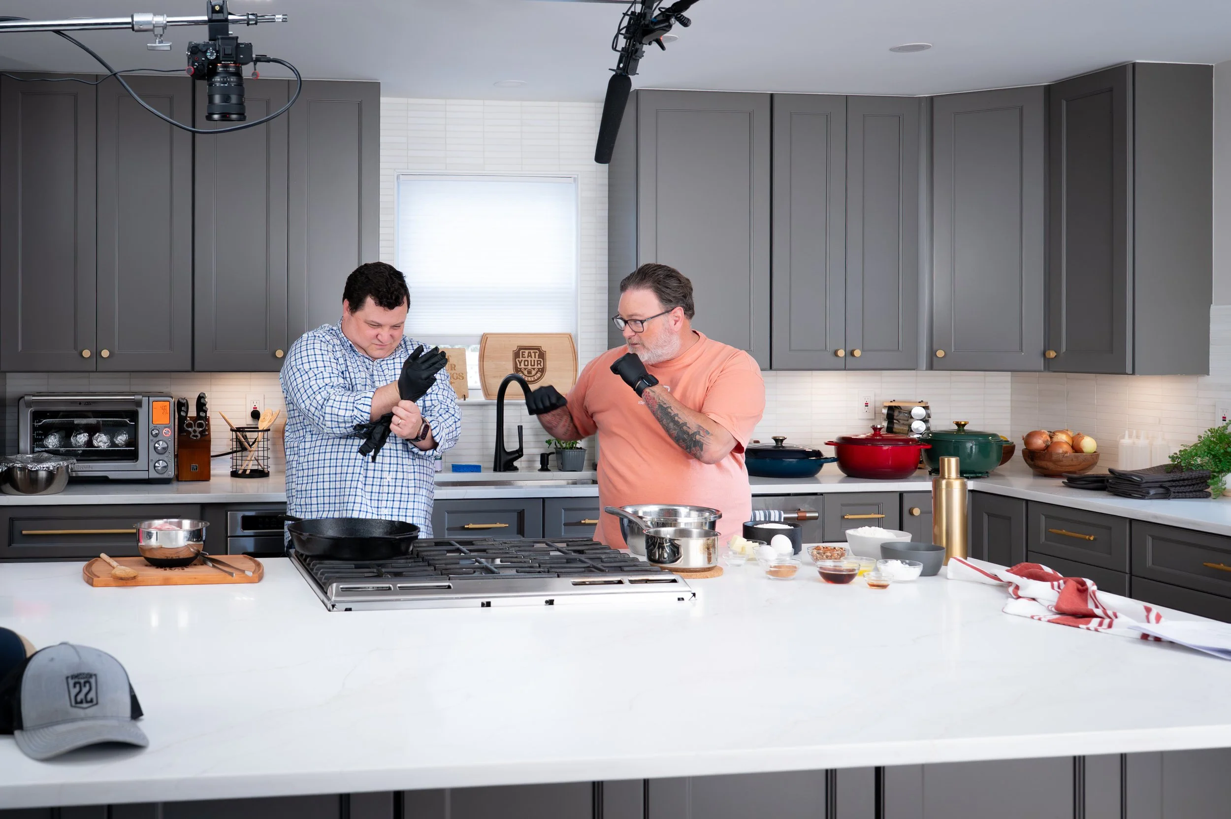 Two men in a modern kitchen, one young and one older, preparing for a cooking segment. The younger man is putting on black gloves, and the older man is inspecting something in his hand. The countertop is scattered with cooking ingredients and utensils.
