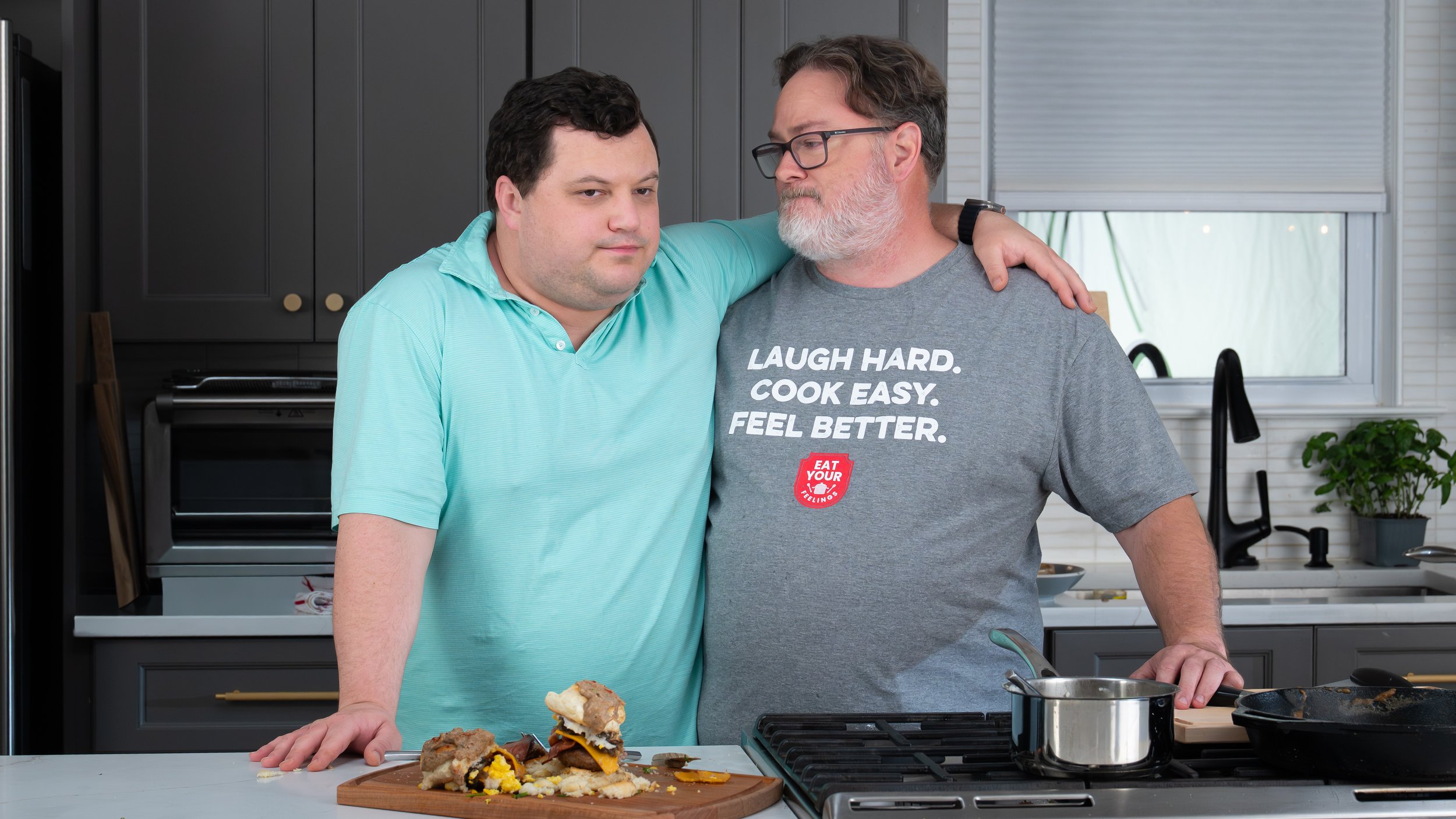 Two men in a kitchen, one with his arm around the other's shoulder, standing next to a counter with food and cooking utensils.