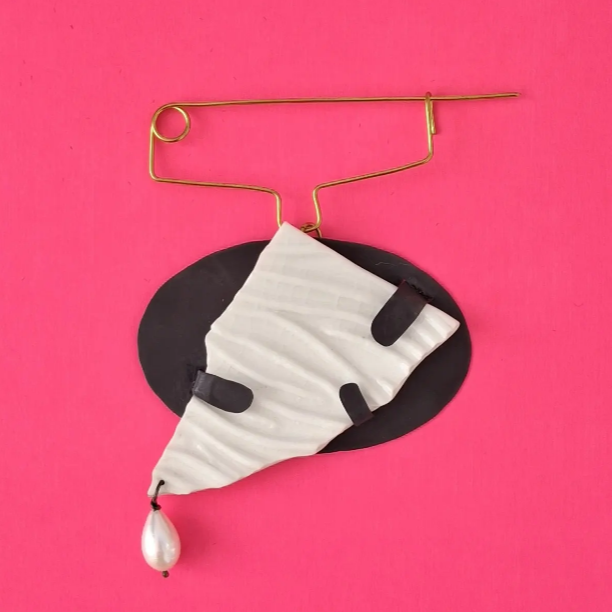 Fashion accessory display with a black oval tag, a white face mask with black accents, a large faux pearl, and a gold safety pin, against a pink background.