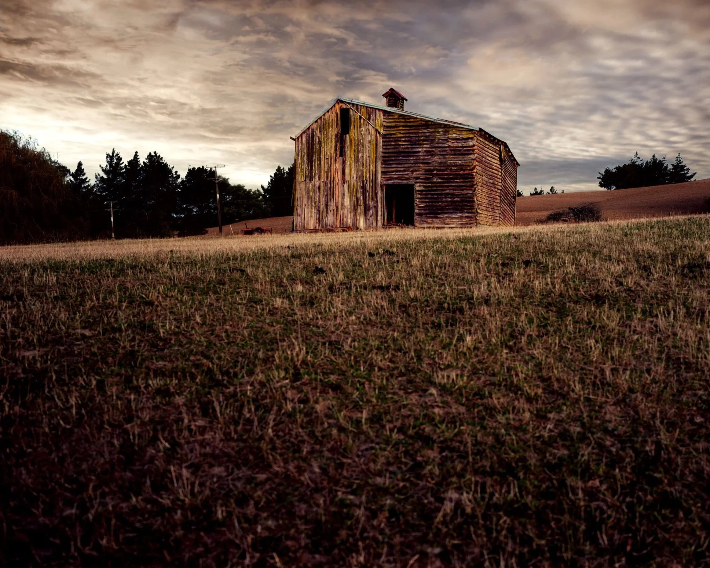 An old weathered barn on a grassy field with trees and utility poles in the background under a cloudy sky