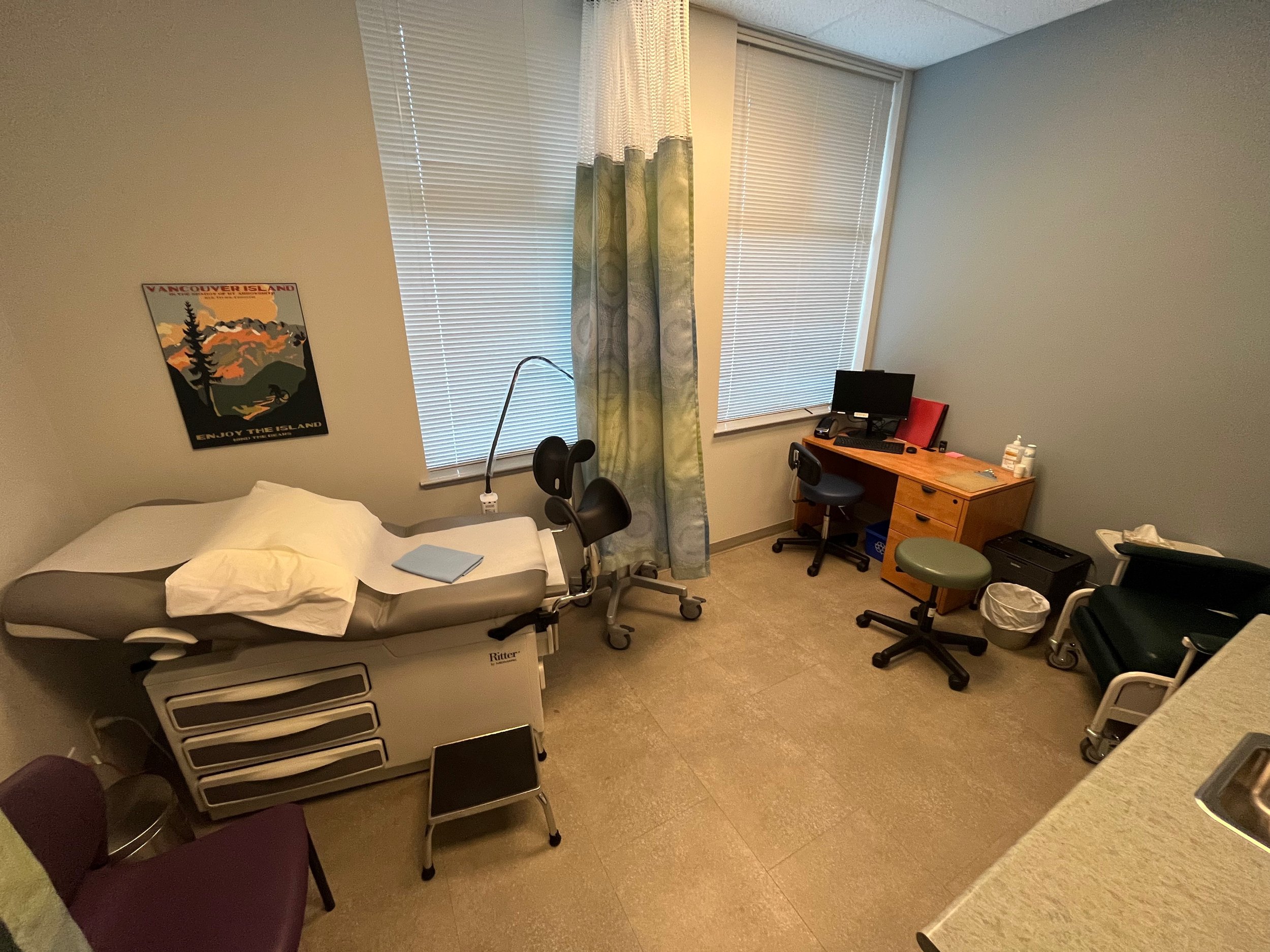 The image shows a medical examination room. On the left, there is an examination bed with a step stool beside it, and a disposable paper cover is placed on the bed. Above the bed, there is a poster on the wall that about Vancouver Island. On the righ