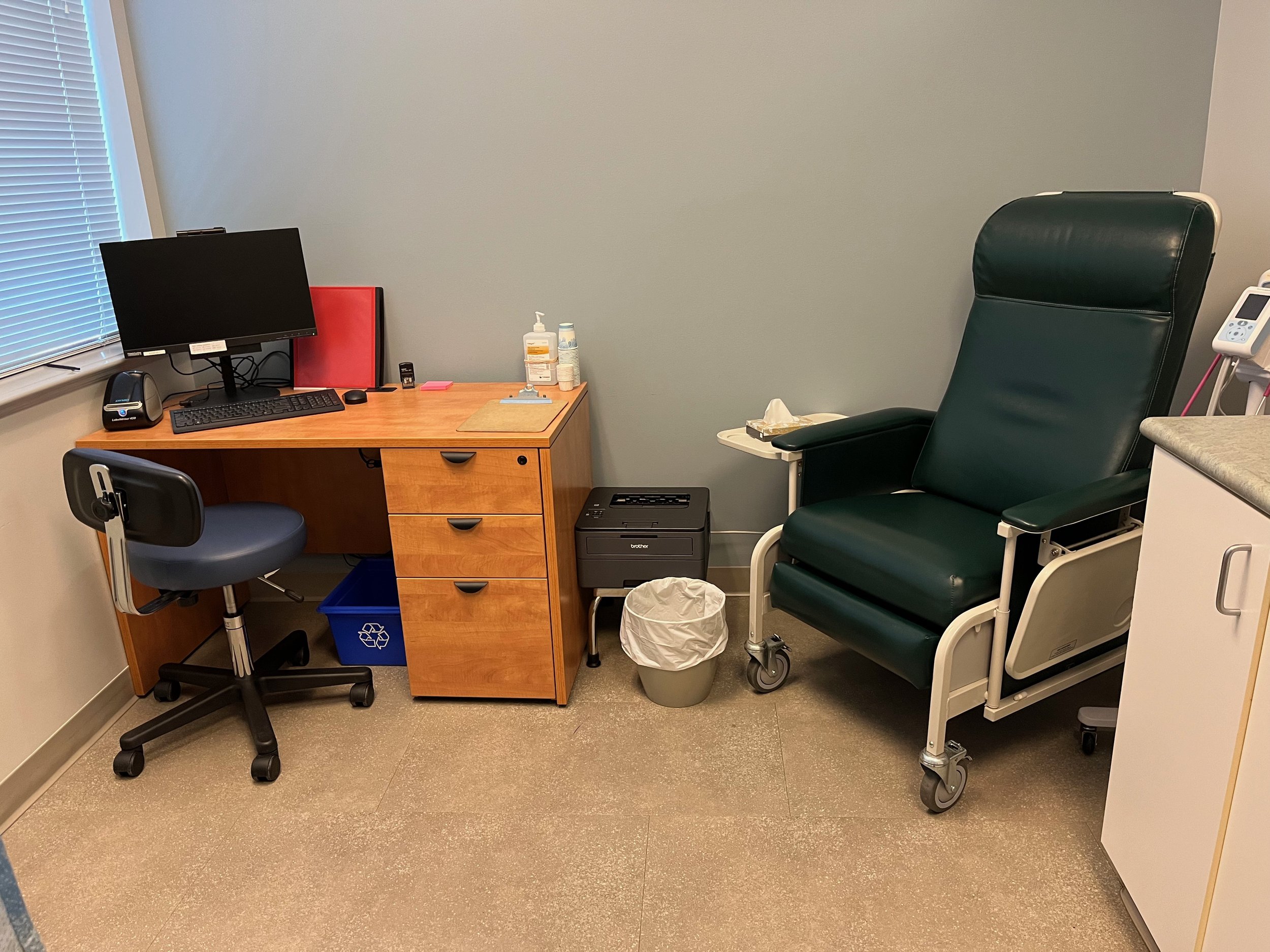This is a medical examination room. On the left, there is a wooden desk with a computer setup including a monitor, keyboard and mouse. There are also some items on the desk such as a bottle of hand sanitizer, sticky notes, and a red folder. An office