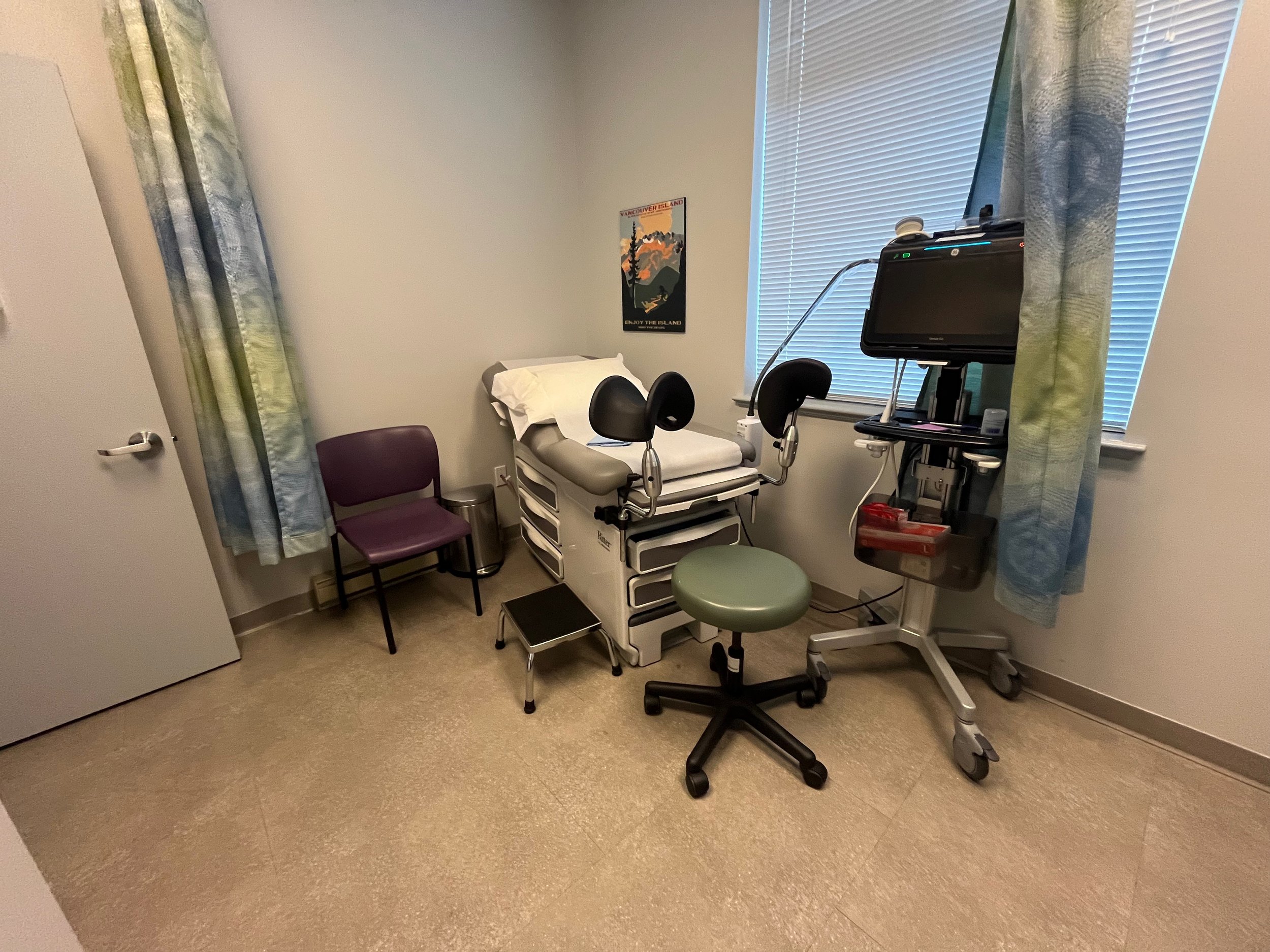 The image shows a medical examination room. There is an examination bed at the center, which is designed for procedures. Attached to the bed are two stirrups to hold a patient's knees in the air to facilitate a pelvic examination. Next to it is a sm