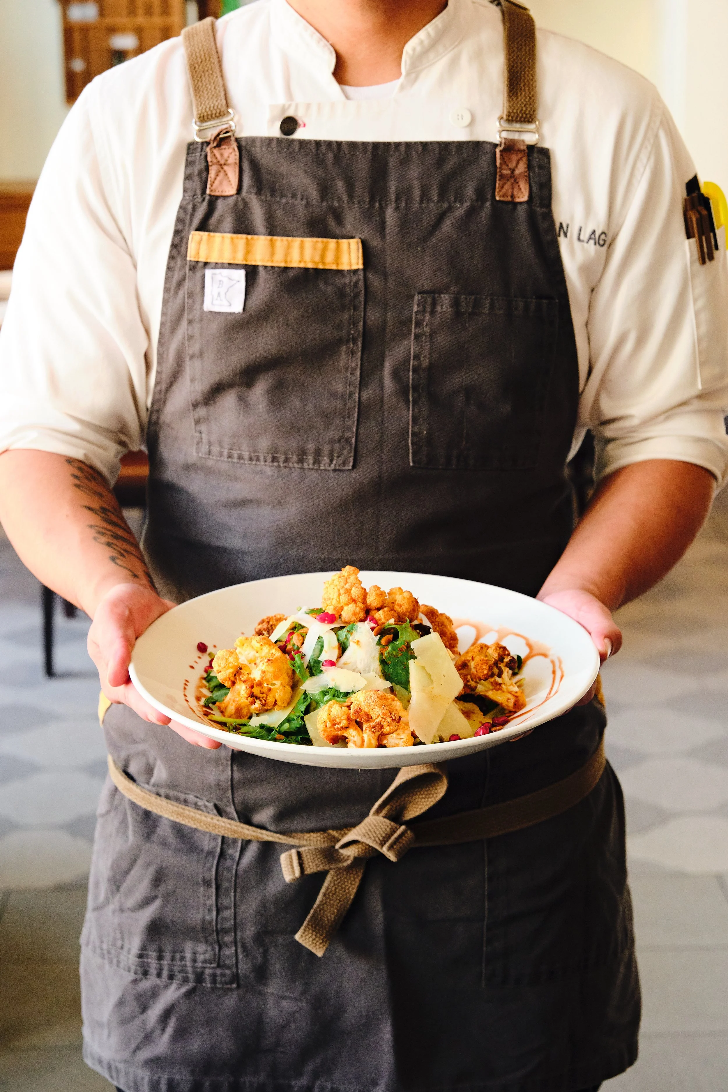 A chef in a gray apron and white shirt holding a plate of Caesar salad with cauliflower, shaved cheese, and pomegranate seeds.