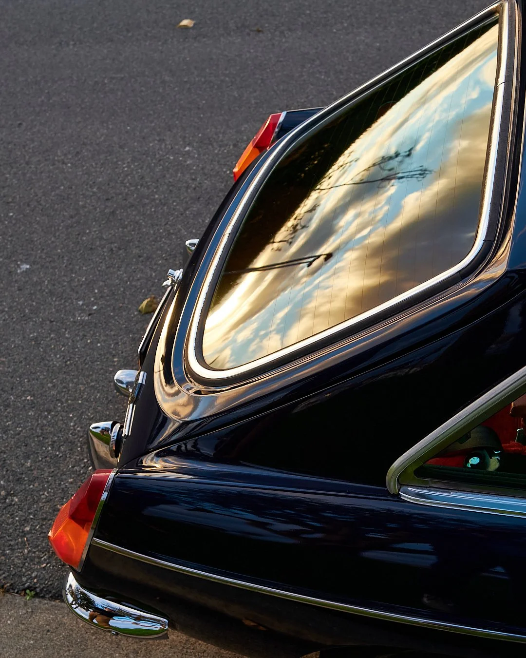 The rear window of a vintage black car reflecting a cloudy sky and power lines, parked on an asphalt surface.