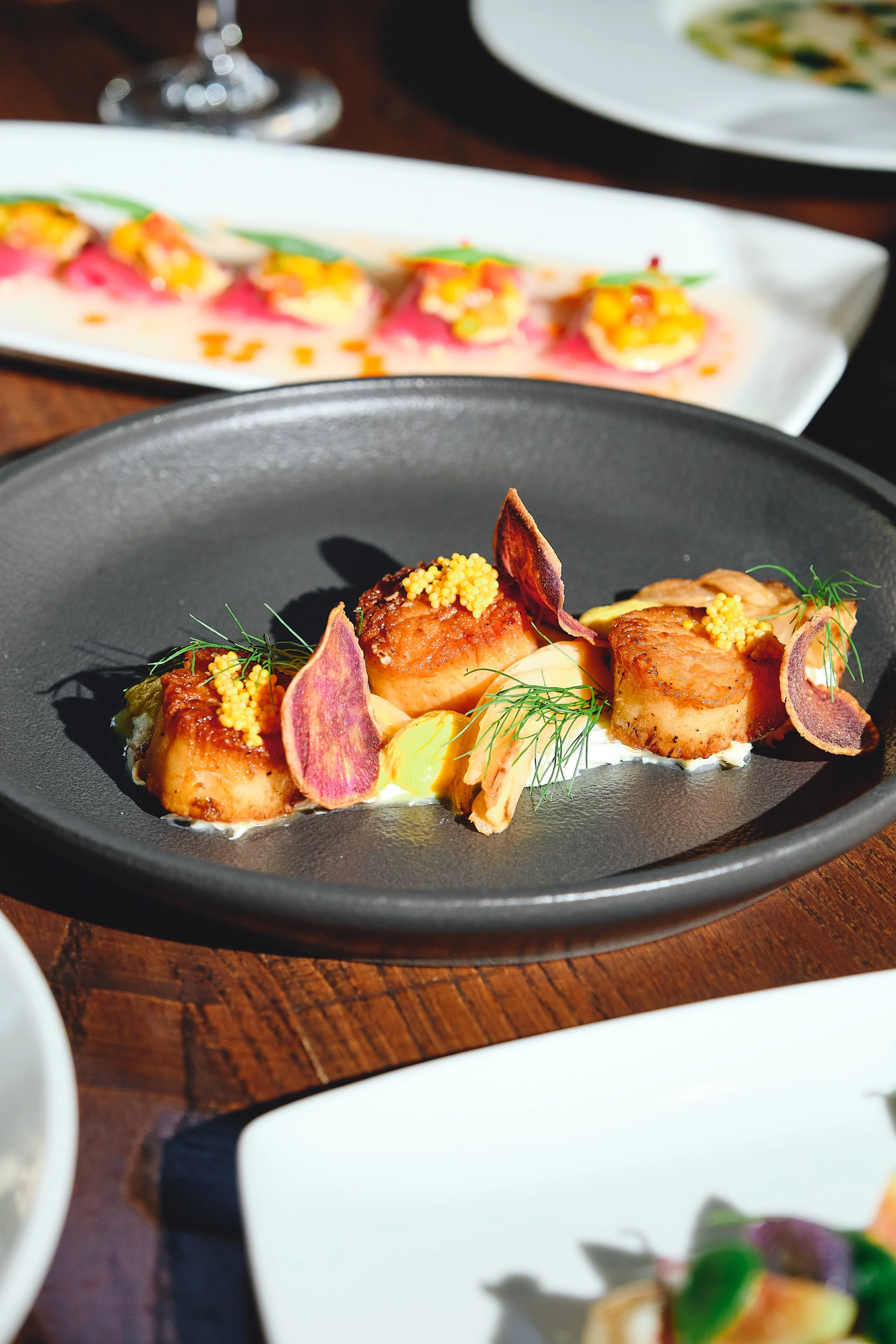 Seared scallops garnished with herbs, edible flowers, and mustard seeds on a black plate.
